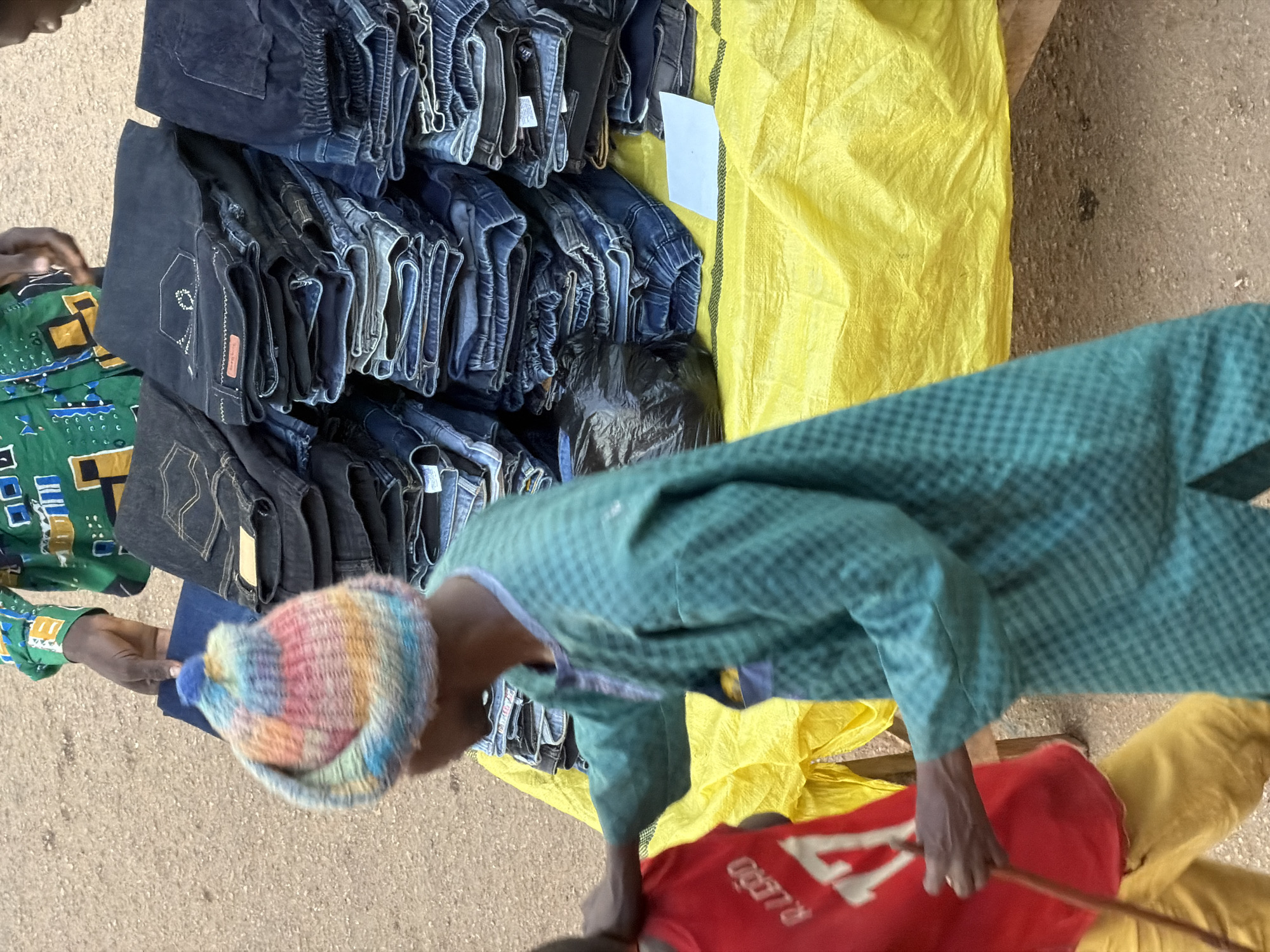 The media file depicts a market scene in Bukuru, Nigeria, likely during daylight hours given the bright lighting. The primary subjects are a group of people interacting with a stall laden with stacked denim jeans. A person in the foreground is wearing a green checkered outfit and a multicolored knitted cap, and appears to be examining or handling a pair of jeans. Another individual, partially visible to the left, wears a green patterned shirt.
The objects on display are predominantly dark blue denim jeans, neatly folded and stacked on a yellow tarp. Some jeans have visible tags and stitching details. A black plastic bag is also visible amongst the clothing. The ground appears to be dirt or gravel.
The activity suggests a commercial transaction, with people browsing or purchasing clothing. There are no strong emotional cues evident, but the setting is a typical outdoor market environment. The image is taken from a slightly elevated, downward-facing perspective. No specific time of day or weather conditions are discernible beyond the general brightness. The location context of Bukuru, Nigeria, helps frame the cultural and economic setting of the scene.