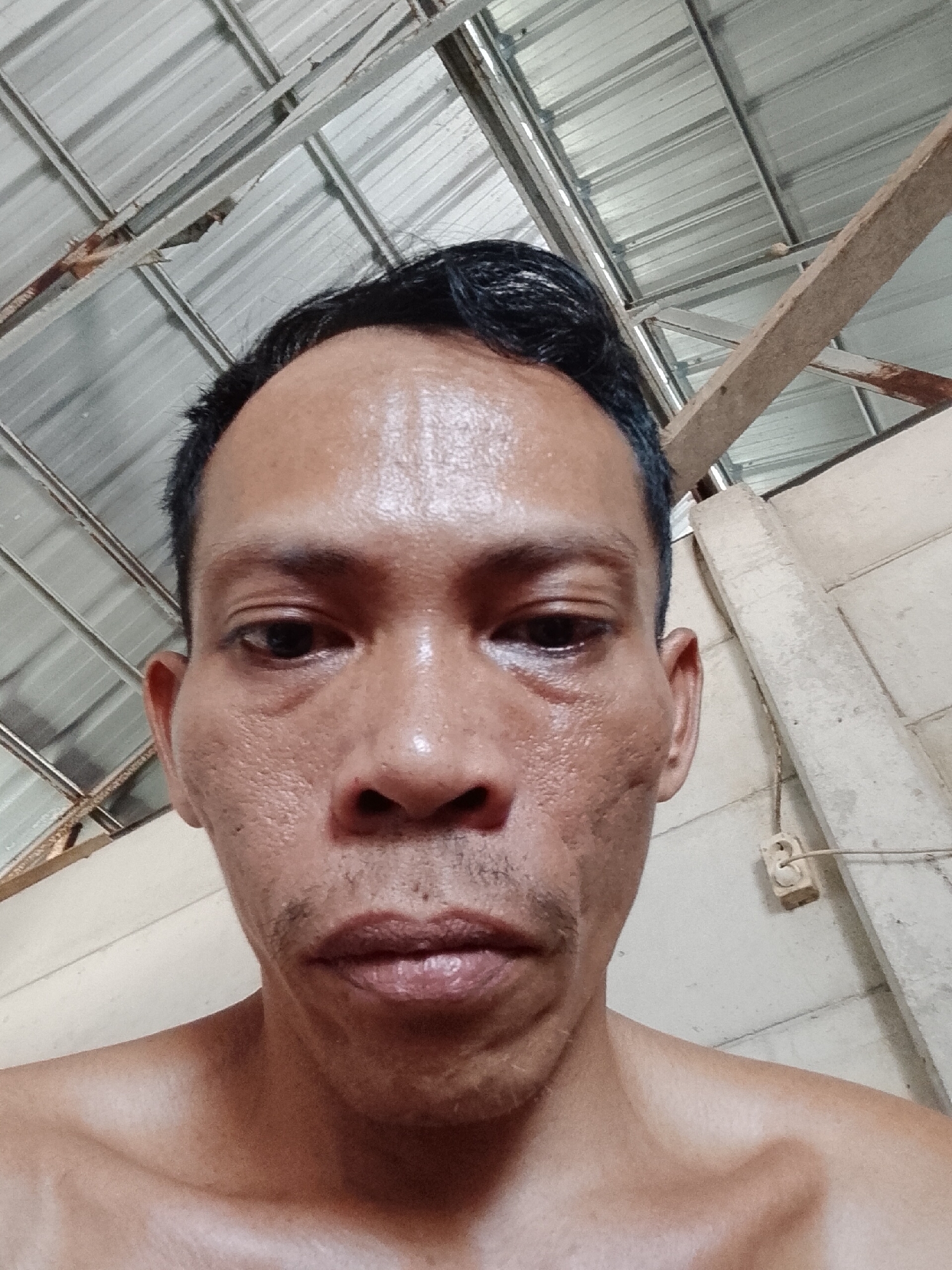 A close-up, upward-angled selfie of a middle-aged Southeast Asian man with short, dark, slicked-back hair. His face is shiny, possibly from sweat or oil, and he has a thin mustache and stubble. The background includes a metal corrugated roof, wooden beams, and a light-colored wall with an electrical outlet. Location is assumed to be Bukit Ketok, Indonesia.
