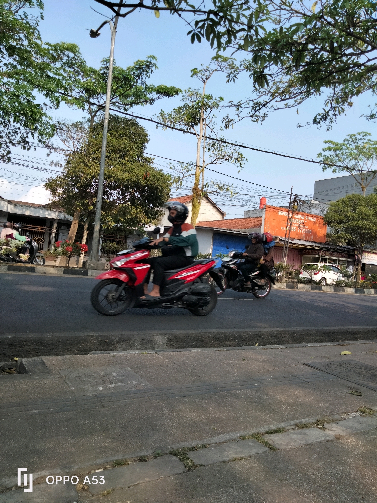 An eye-level, medium shot captures a street scene in Kelutan, Indonesia, under a clear blue sky, with dappled sunlight filtering through overhead trees and utility wires crisscrossing the background. The foreground features a concrete curb and sidewalk, with some sparse vegetation. The primary action centers on a vibrant red scooter, with a rider wearing a helmet and a dark jacket with colored accents, moving from left to right across the asphalt road. To the right of the red scooter, another scooter, dark in color, is also in motion, carrying two individuals, both wearing helmets. In the mid-ground, buildings line the street. The building directly behind the scooters is orange with "KOPERASI SIMPAN PINJAM SAHABAT" signage and a phone number. Further down the street, a white car is visible. The overall impression is a typical urban environment in Indonesia during daylight hours, with a focus on vehicular traffic.