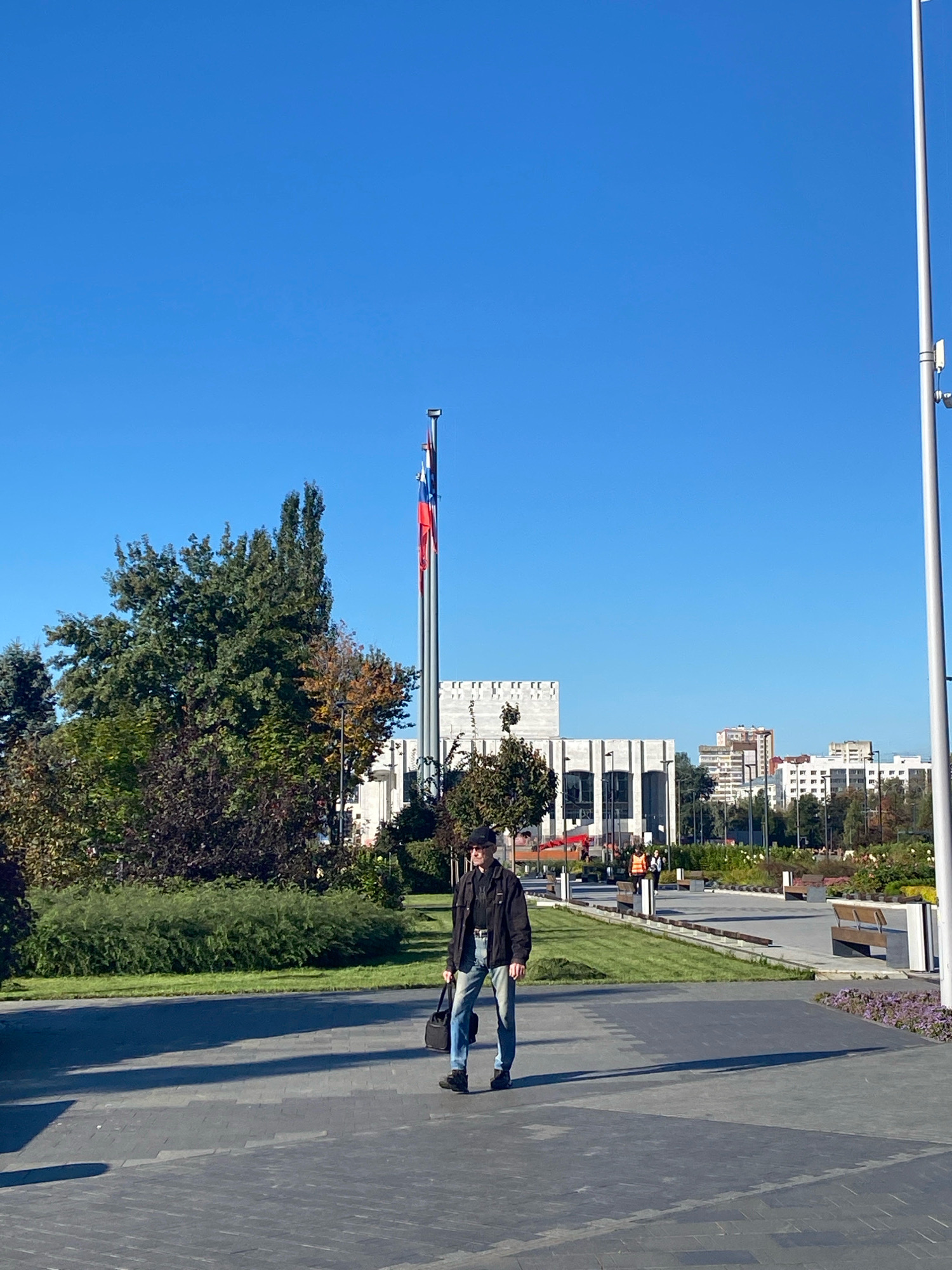 A lone man wearing a black jacket, black hat, and light blue jeans walks from right to left across a paved plaza on a sunny day. He is carrying a black messenger bag in his right hand. In the background, a large, modern, white building with vertical lines dominates the scene. Several flags, including the Russian flag, fly on tall poles in front of the building. Trees with green and autumn-colored foliage are to the left of the building, and a well-maintained lawn is in front of them. The plaza features dark gray paving stones with lighter gray patterns. Benches are visible further back in the plaza, and in the distance, several other buildings, including apartment complexes, can be seen. The sky is a clear, vibrant blue.