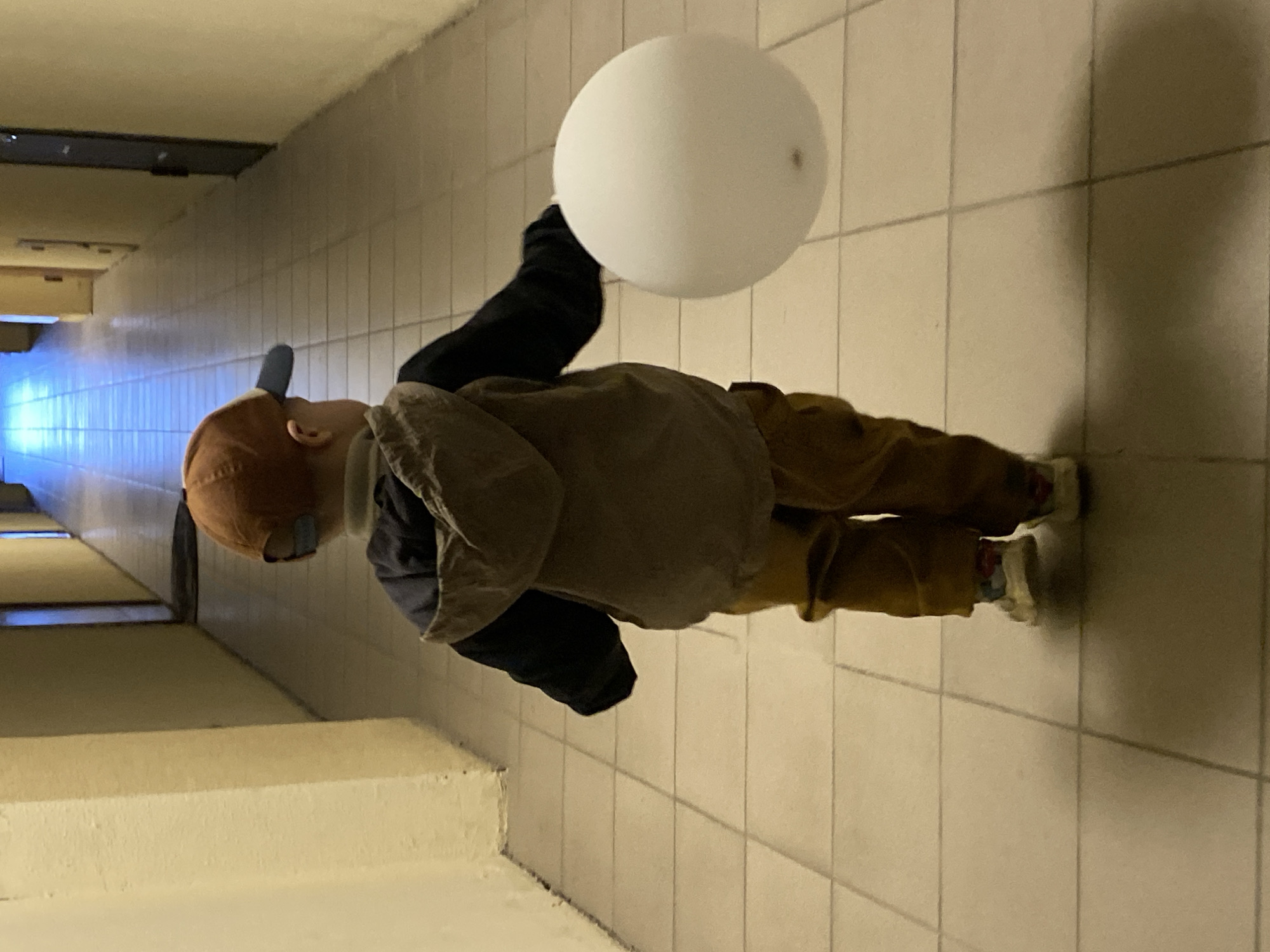 A young child stands in a tiled hallway, holding a white balloon. The hallway has a series of doors visible on the left side, leading into the distance. The child wears a brown jacket, dark pants, and a cap. The floor tiles are light-colored and the hallway appears to be indoors. The location within Sochi, Russia is not discernible from the provided image.