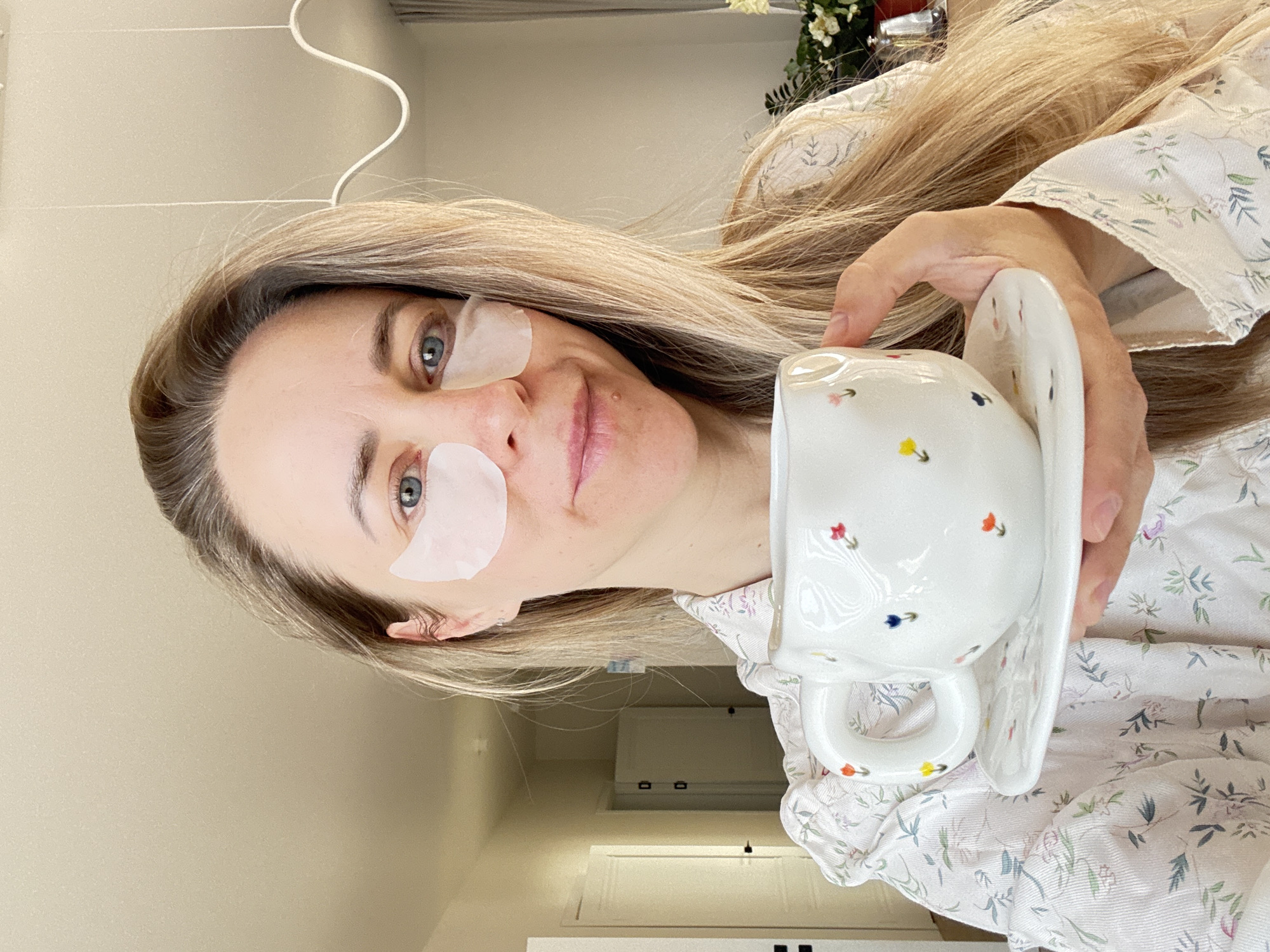 A woman is indoors, wearing eye masks and holding a white floral-patterned teacup and saucer. Her hair is blonde, and she is wearing a floral patterned top. A white door and part of the ceiling are visible in the background. The location appears to be indoors, and it cannot be confirmed as being in the city of Chaikovsky, Russia, from the given image.