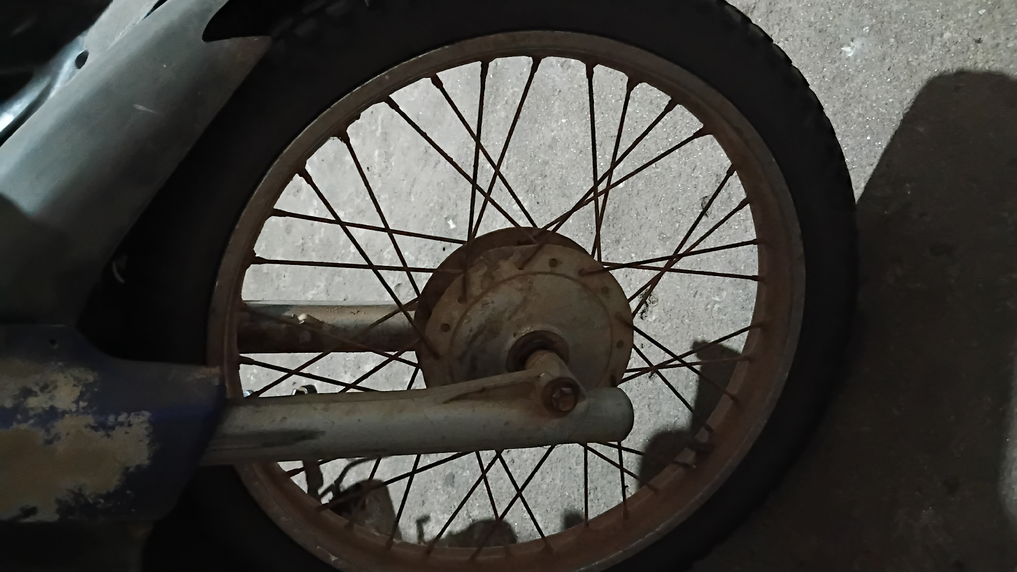 The image shows the front wheel of a motorcycle, focusing on the spokes, rim, and tire. The motorcycle's front fork and a portion of the fender are also visible. The scene appears to be outdoors, possibly on a street or parking area, with a concrete or asphalt ground. The overall condition of the motorcycle part suggests it is old or has been exposed to the elements, as indicated by rust on the spokes and rim, and peeling paint on the fender. There are no people or any visible signs of activity in the image. The lighting suggests it might be evening or night, with shadows cast on the ground. The image does not provide any specific details that would allow for precise location identification within Prabumulih, Indonesia; it depicts a common urban scene.