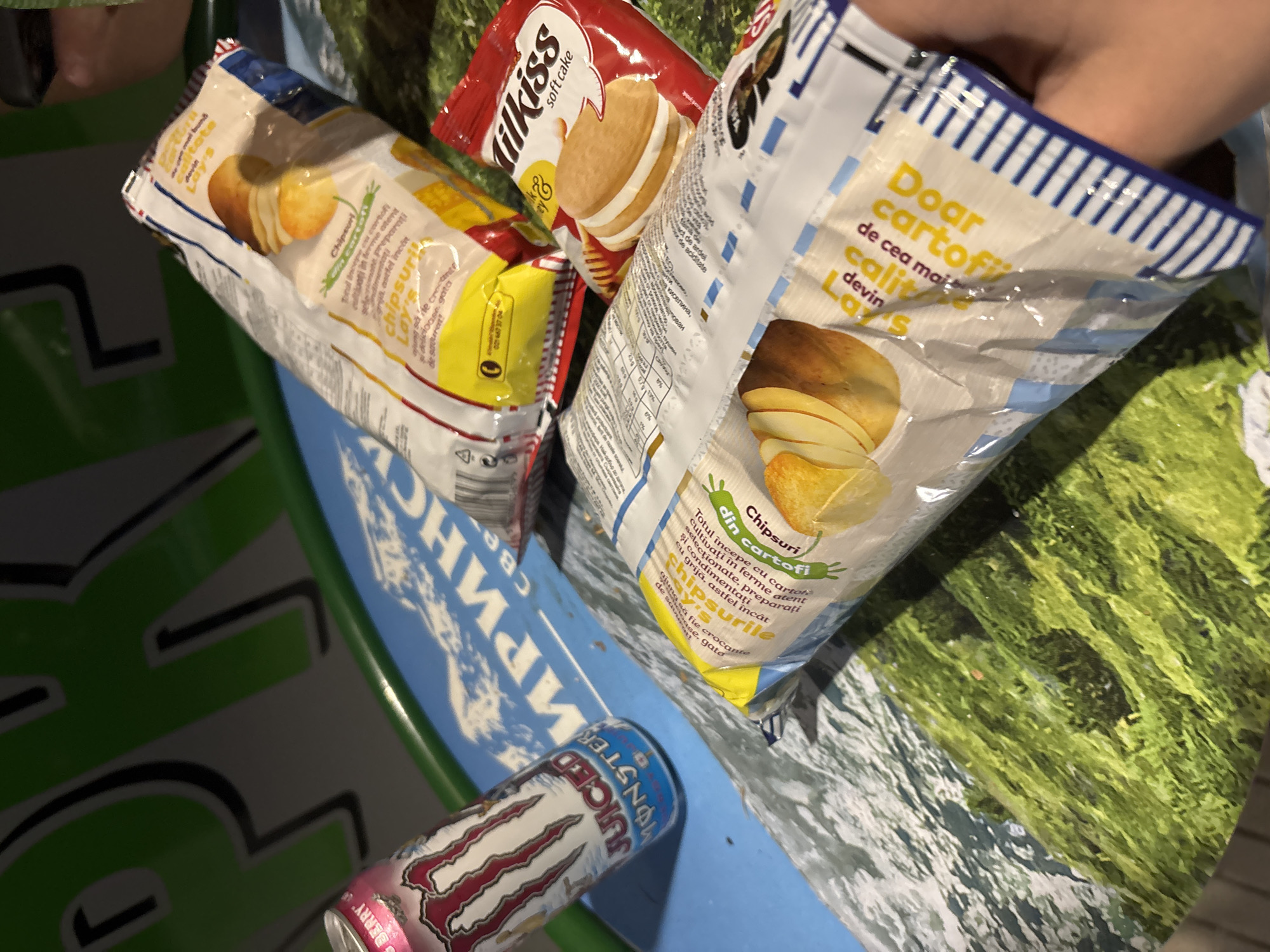 This close-up, high-angle shot captures a collection of snack items and a beverage can in what appears to be a shopping basket or cart, likely in Sandanski, Bulgaria. A hand is visible reaching into the basket, possibly selecting or arranging items.
The main subjects are three bags of snacks and one energy drink can. Two of the snack bags are Lay's potato chips, featuring Romanian text such as "Chipsuri din cartofi" (Chips from potatoes) and descriptions like "Totul începe cu cartofi selectționate, cultivați în ferme atent" (Everything starts with selected potatoes, carefully cultivated) and "Doar cartofi de cea mai înaltă calitate devin Lay's" (Only the highest quality potatoes become Lay's). One Lay's bag mentions "ardei" (pepper) and "aciditate" (acidity), indicating a flavor. Another snack is a red bag of "Milkiss soft cake" with a depiction of a cream-filled cookie. The beverage is a pink and silver Monster Energy drink can, specifically "JUICED MONSTER", with "BERRY" visible at the bottom, and a partial Cyrillic inscription "МАРИНСV" possibly part of a store or promotional detail.
The background of the basket/cart features a stylized landscape print with green areas resembling grass or trees and blue and white patterns suggesting water or sky. The overall impression is a casual scene of a shopping trip or recent purchase. The time of day is not discernible, but the setting is indoors.