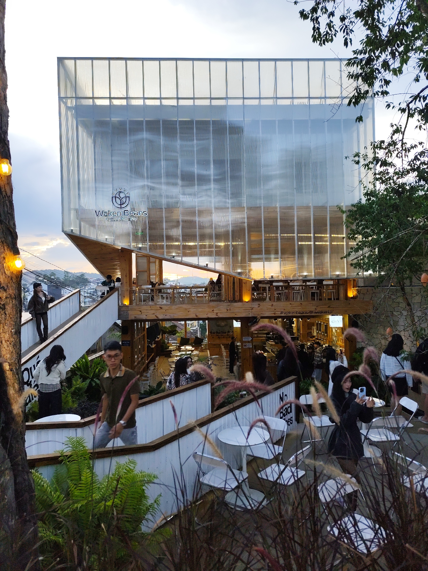 The image depicts a multi-level outdoor café named "Waken Beans" located in Da Lat, Vietnam. The café is built into a hillside with a modern architectural style, featuring a translucent exterior and wooden support beams. People are seen on different levels of the café, some walking up stairs, others seated at tables. The café includes outdoor seating areas with white tables and chairs. The scene is partially obscured by tall grasses and plants in the foreground. The lighting suggests it is late afternoon or early evening.