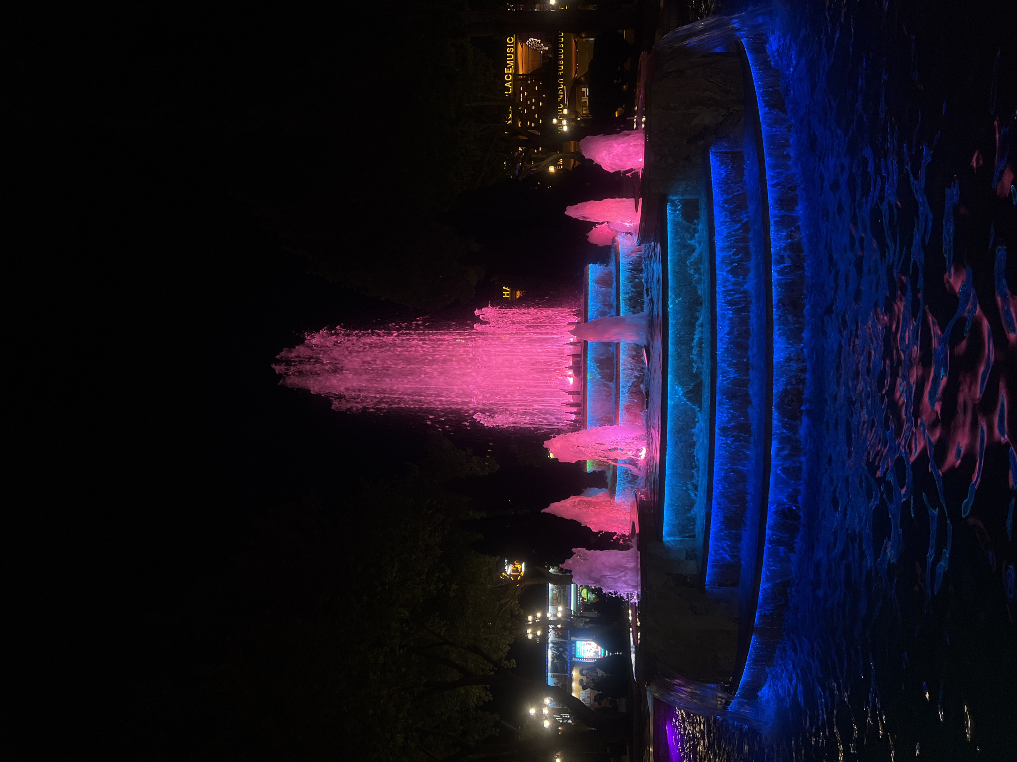 The image captures a nighttime scene of a brightly illuminated fountain in Yerevan, Armenia. The fountain features a central jet of water lit in pink, along with smaller jets of the same color cascading down stepped platforms. The platforms themselves are illuminated with blue light.
In the background, blurred lights and signs indicate the presence of nearby buildings and possibly amusement attractions; one sign reads "PLACE MUSIC."
The foreground shows water flowing from the illuminated fountain, reflecting both the pink and blue lights. The composition suggests a long exposure, as the water's movement creates a blurry effect.