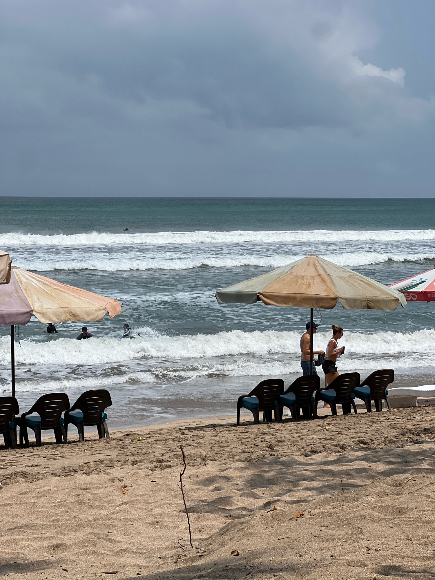 A frontal, medium shot shows a beach scene with people swimming in the ocean and sunbathing on the sand, with a stormy sky overhead. The beach is located in Kuta, Indonesia, a popular tourist destination known for its surfing and lively atmosphere. In the foreground, several empty beach chairs are arranged on the sand, facing the ocean. Two umbrellas are set up, one in the left foreground and another in the center, providing shade.
In the middle ground, a few people are seen wading and swimming in the choppy ocean waves. A couple stands under the central umbrella, seemingly conversing. The water is a deep blue-green, with white foam from the breaking waves. The sky is overcast with dark, heavy clouds, suggesting an impending storm. The sand is a light brown color, with some scattered debris and leaves. The overall mood is one of anticipation, as the weather suggests a shift.