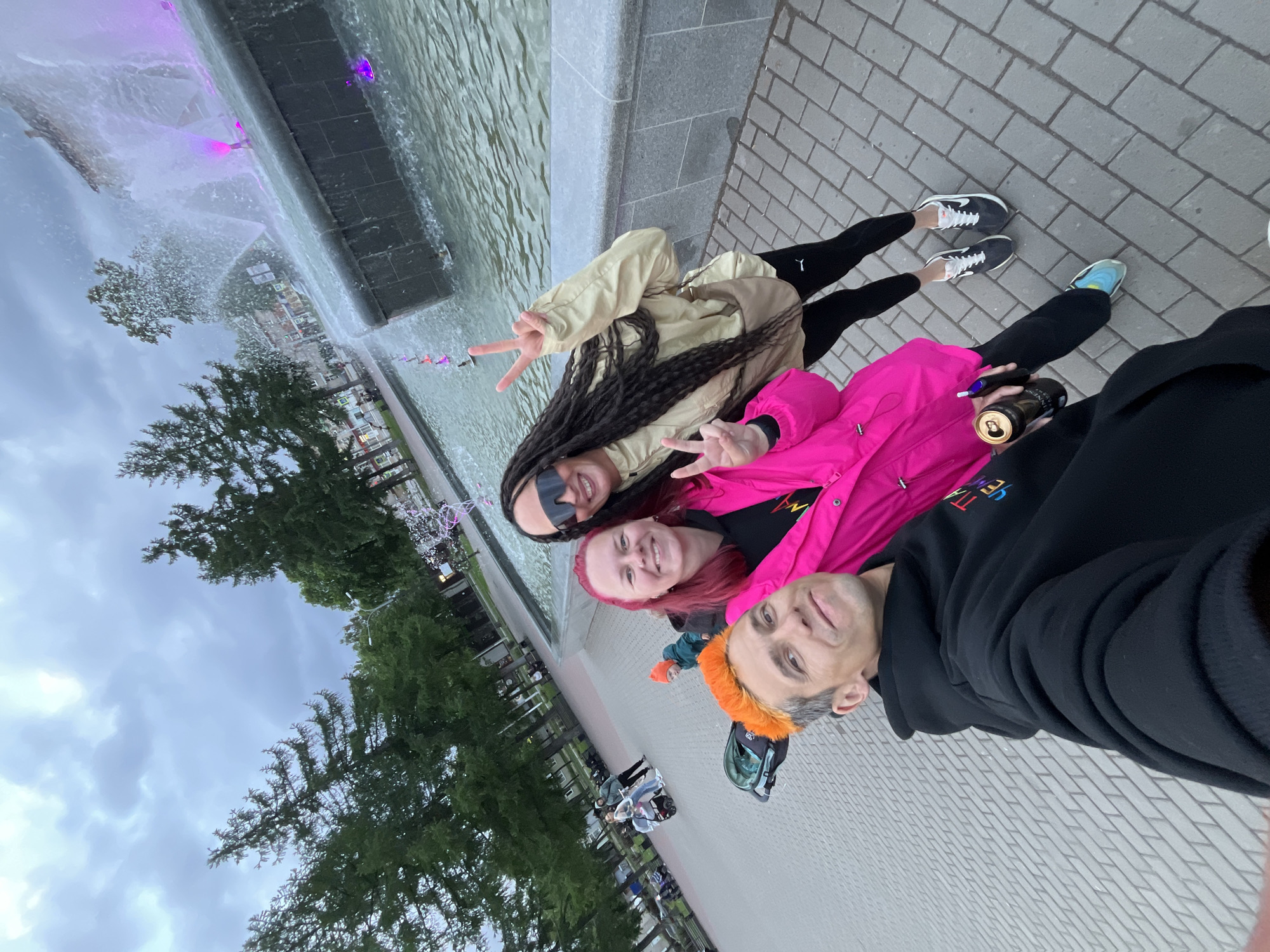 The image displays a selfie of three individuals in a public park or square in Chaikovsky, Russia, under an overcast sky.
In the foreground, from left to right:
1. A male individual with short grey hair and prominent orange dye on top of his head. He is wearing a black hoodie with partially visible colorful Cyrillic text on the front. He exhibits a slight smile and holds a dark-colored can in his right hand. His left arm extends forward, presumably holding the camera.
2. A female individual with reddish-pink hair and fair complexion, wearing a bright pink jacket over a dark top. She smiles broadly, presenting a 'peace' sign (V-sign) with her right hand near her face. Her left hand holds a black cylindrical object, possibly a vape pen, and a dark-colored can. She wears a multi-colored sneaker (blue and green tones) on her left foot.
3. A female individual with dark skin, long dark dreadlocks, and large dark sunglasses. She is wearing a beige/khaki jacket, black leggings with a white 'Puma' logo on the left thigh, and black-and-white sneakers. She also smiles broadly and displays a 'peace' sign with her right hand. A beige crossbody bag is slung across her body.
The midground features a grey brick-paved ground extending into the distance. To the left, numerous tall coniferous trees, possibly spruces or firs, with dark green foliage are visible, interspersed with some deciduous trees. Several individuals are present in the background: one person is pushing a light-colored baby stroller, another individual in dark attire stands nearby, and a child wearing an orange hat and a green jacket is visible behind the central foreground figures. A grey and black object, possibly a child seat or stroller component, rests on the ground near the male's feet.
To the right, a large, multi-tiered dark grey stone fountain actively sprays water. The water within the fountain is illuminated by purple and pink lights emanating from beneath the surface. Beyond the fountain and trees, distant buildings with light-colored facades are partially visible.
The sky is uniformly overcast with grey clouds, providing diffused and even lighting across the scene.