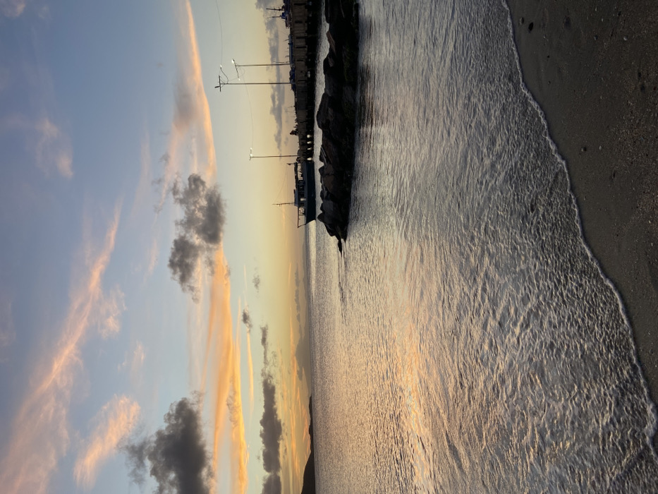 A tranquil beach scene bathed in warm sunlight. The ocean glistens with subtle ripples, leading to a sandy shore on the right. To the left, a pier or breakwater stretches into the water, with a boat docked alongside. Above, a sky painted with hues of blue, orange, and yellow reflects the sunset, punctuated by scattered clouds that mirror the golden light. The composition evokes a sense of peace and natural beauty.