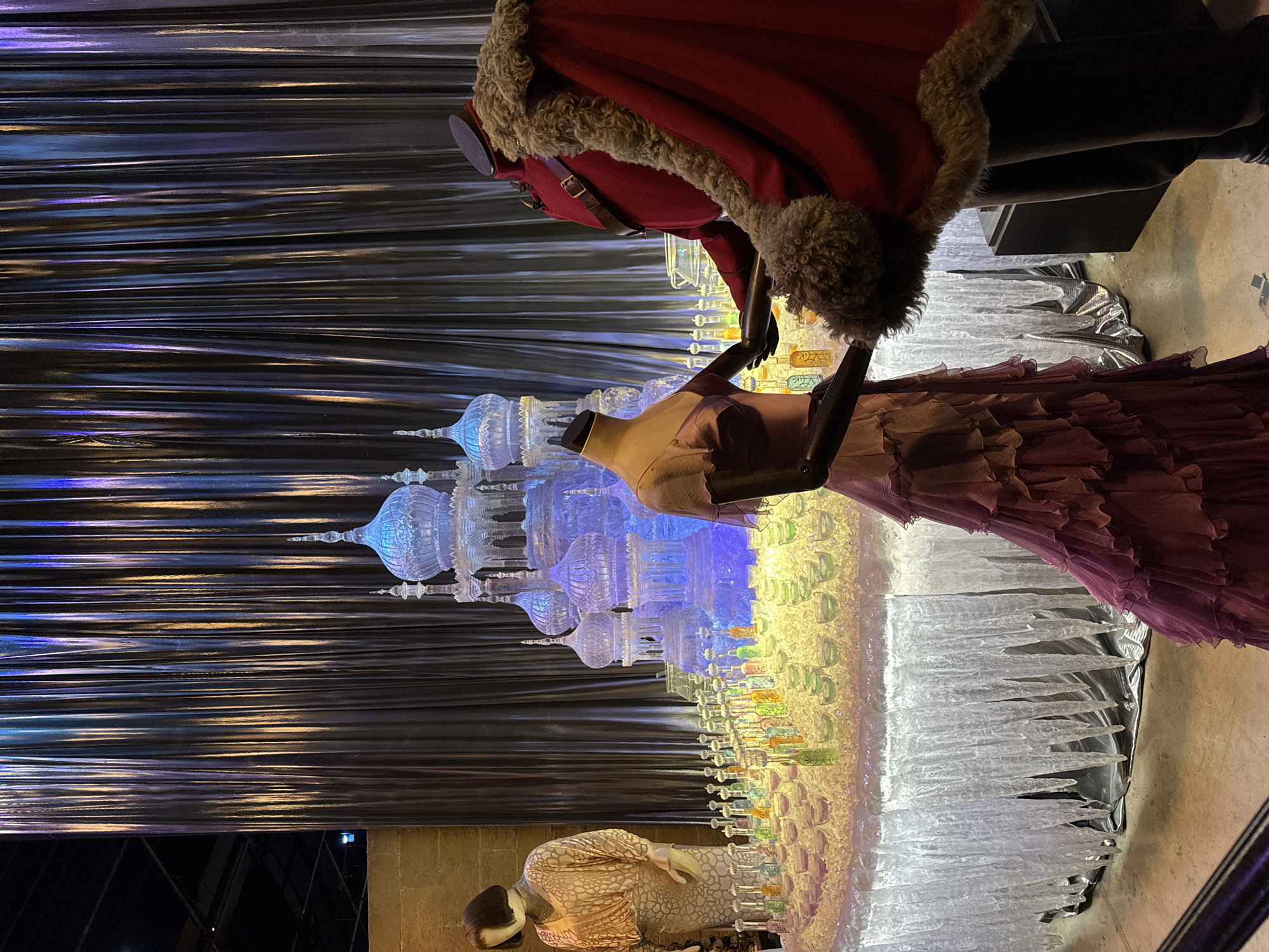 The image depicts a display, likely from a film set or exhibition, at Warner Bros. Studio Tour London – The Making of Harry Potter, located in Watford, United Kingdom. In the foreground, two mannequins are positioned. The mannequin on the left wears a light-colored, patterned dress. The mannequin on the right is dressed in a red coat with a fur collar, a dark belt, and dark trousers.
The central focus of the scene is a large, elaborate ice sculpture shaped like a castle with multiple domes and spires, illuminated with blue light. In front of the ice sculpture is a table covered in crushed ice, upon which are arranged numerous small bottles filled with colorful liquids, presumably potions, and many small glasses. The table itself is draped with a white, icicle-like material.
Behind the ice sculpture, a silver, pleated curtain forms a backdrop, reflecting the light. To the left of the ice sculpture and mannequins, another mannequin is visible, wearing a cream-colored dress with an intricate pattern. The environment appears to be indoors, with a concrete floor visible in the lower portion of the image. The overall impression is one of a fantasy or festive setting.