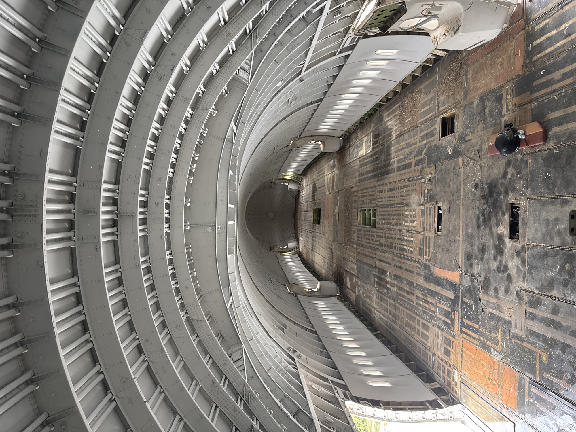 The image shows the interior of a disused airplane fuselage, likely a Boeing 747, with its interior stripped bare. The ribbed, grey metal structure of the ceiling and walls is visible, with rows of oval windows lining the sides. The floor is made of dark, worn wood panels with visible markings and some orange discoloration, suggesting previous use or damage. There are several access hatches in the floor. At the far end of the fuselage, the circular shape of a bulkhea is visible. In the foreground, on the floor, lies a black helmet on a red and beige object. The scene appears to be located in an open, possibly outdoor, environment given the natural light and greenery visible through the open doors on the left side. The location is identified as being in Pak Nam Pran, Thailand. There are no people visible in the image.