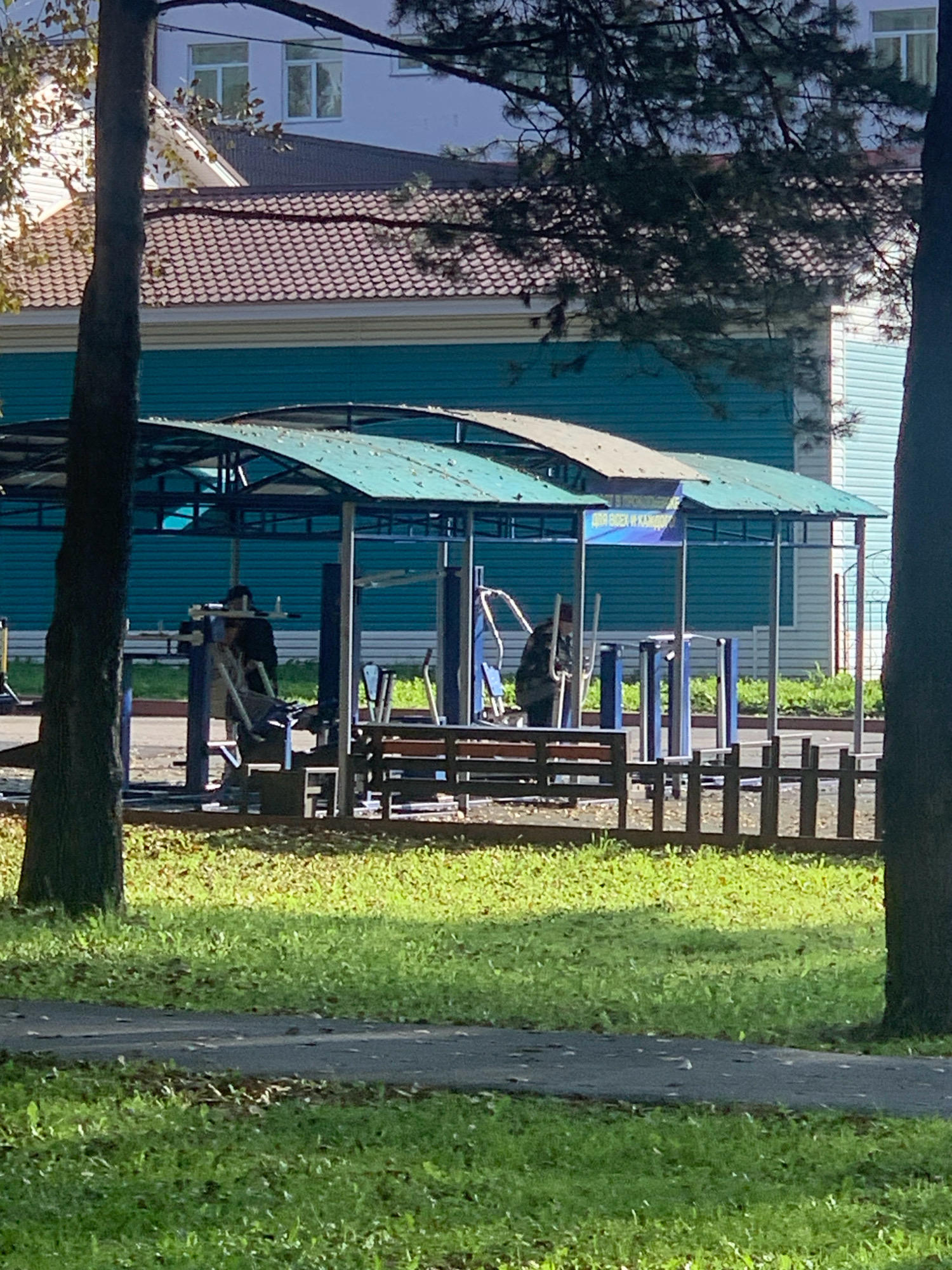 An outdoor exercise area in Prokopyevsk, Russia, features two individuals engaged with workout equipment beneath a covered structure. The area is surrounded by a wooden fence and green grass. In the background, a teal-colored building with white trim and windows is visible, along with a dark-tiled roof. Trees with green and brown foliage frame the scene, and a paved path runs through the grass.