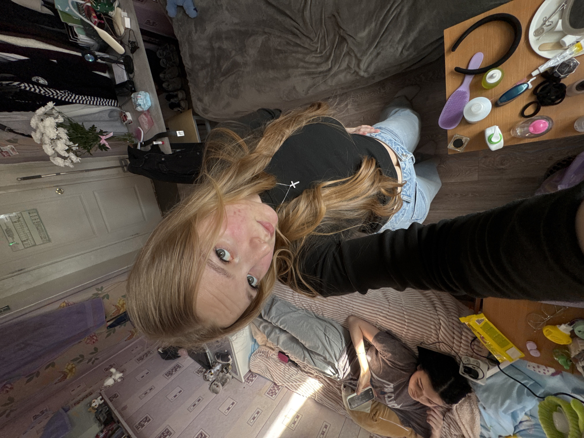 A high-angle selfie captures two individuals within a cluttered indoor setting. In the foreground, a person with long blonde hair, wearing a black long-sleeved top and light blue overalls, looks directly at the camera with a neutral expression. A silver cross necklace adorns their neck.
In the background, a second person with short dark hair lies on a bed with striped and grey bedding, facing away from the camera, seemingly resting or using a device. Natural light streams into the room, illuminating parts of the scene and suggesting it is daytime.
The room appears to be a bedroom or living area, featuring a grey textured couch, wooden flooring, and wallpapered walls (one with a floral pattern, another with a subtle geometric design). Numerous personal items are visible: a bouquet of white chrysanthemums with a pink accent flower rests on a surface next to piles of clothes (black, white striped, grey). On a wooden table, a purple hairbrush, black headband, various cosmetic containers, a toothbrush, hair ties, and a small dish with medical tools are scattered. Other visible items include a package of diapers or incontinence pads, a pink card-like item, a silver laptop/tablet with a small sticker, a power strip, a yellow packaged item, and small stuffed animals. The overall impression is a lived-in and somewhat disheveled domestic space.