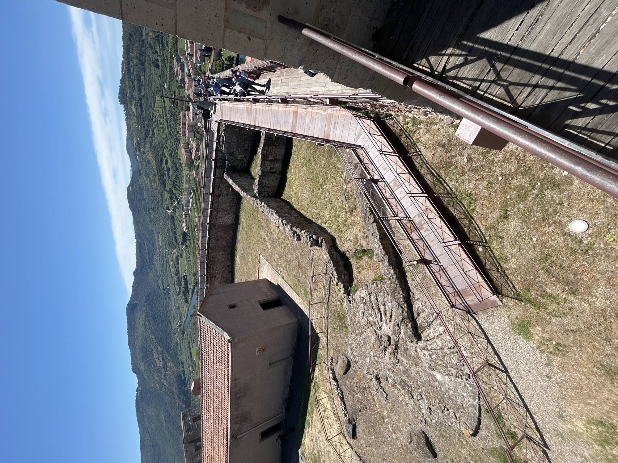 This image, captured from a high vantage point, provides a downward and panoramic view of a historical or archaeological site, likely a castle or fortress in Bosa, Italy, under bright daylight.
In the immediate foreground, the edge of an upper structure is visible, featuring dark wooden planks overhead that cast strong shadows, and two rusty metal pipes or railings extending diagonally.
The mid-ground showcases a complex of ancient stone walls, partially ruined, outlining various sections of the site. A modern, multi-level walkway system, constructed from metal grates and railings, winds through these ruins, indicating visitor access. A group of people, presumed to be tourists, are visible on an elevated section of this walkway in the upper-middle right, exploring the site. Below, a small, light-brown, rectangular building with a red-tiled gabled roof stands within the complex, featuring simple windows or openings. The ground is a mix of dry grass and gravel.
The background unfolds into a scenic landscape under a clear blue sky with a few wispy clouds. Rolling hills, densely covered in green vegetation, dominate the horizon. At the base of these hills, a cluster of buildings belonging to a town is visible, and a slender body of water, likely a river, can be seen winding through the valley.
The scene depicts a sunny day, likely mid-morning or afternoon given the shadow angles, with clear weather conditions suitable for outdoor exploration. No visible text is present in the image.