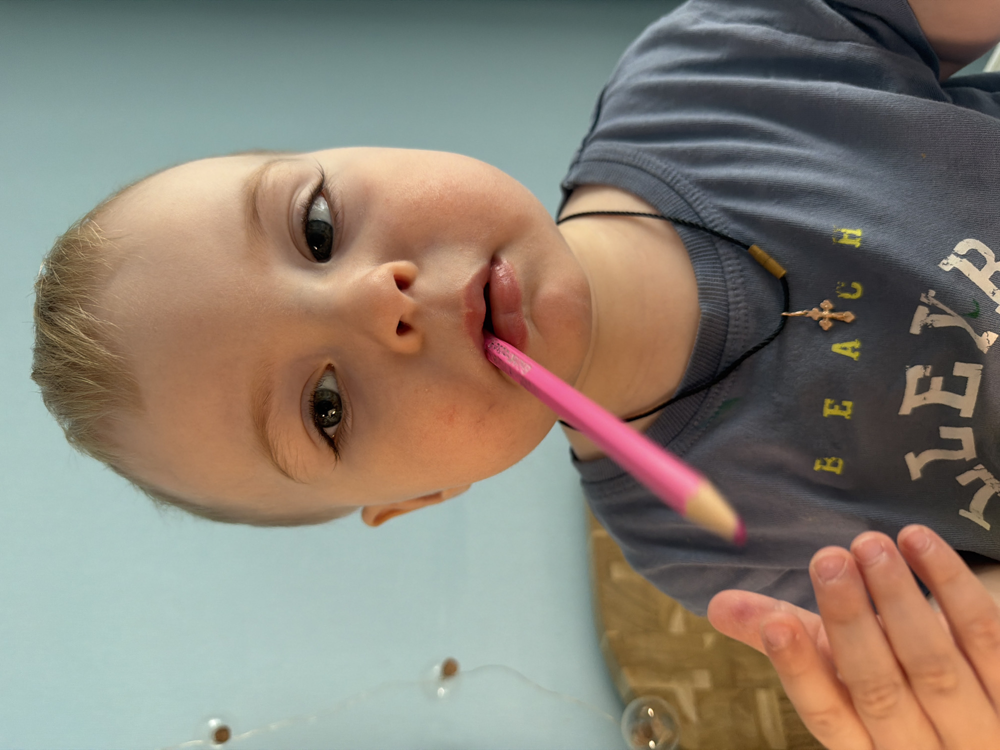 A close-up, angled shot depicts a young child with light skin, light hair, and dark eyes. The child has a pink pencil held in their mouth. They are wearing a gray t-shirt with yellow and white lettering. A black cord with a cross pendant is visible around their neck. The child's right hand is visible in the lower right corner of the frame. The background appears to be a light blue wall or surface. The location is not evident from the image.