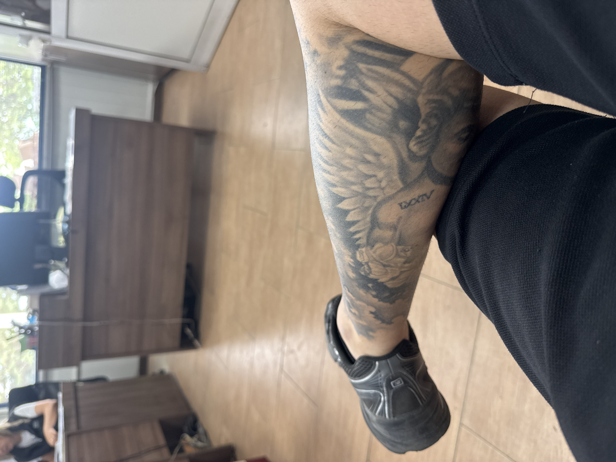 An indoor shot taken in Kovachevo, Bulgaria, shows a person's tattooed arm and leg, highlighting a grayscale tattoo of an angel or winged figure on the forearm. Below the winged figure, flower motifs, possibly roses, are visible. The Roman numerals "LXXIV" are distinctly tattooed on the arm. The person is wearing black casual or athletic clothing, specifically a black top and black shorts or trousers, along with black athletic shoes featuring subtle silver/white accents.
The individual appears to be seated, with their arm bent and resting. The background reveals an office or workspace setting with light brown tiled flooring and dark wooden desks and office chairs. Natural daylight streams in from a window on the left, suggesting it is daytime. In the blurry background, another person can be partially seen seated at a desk, seemingly working. The overall scene conveys a casual moment within an office environment.