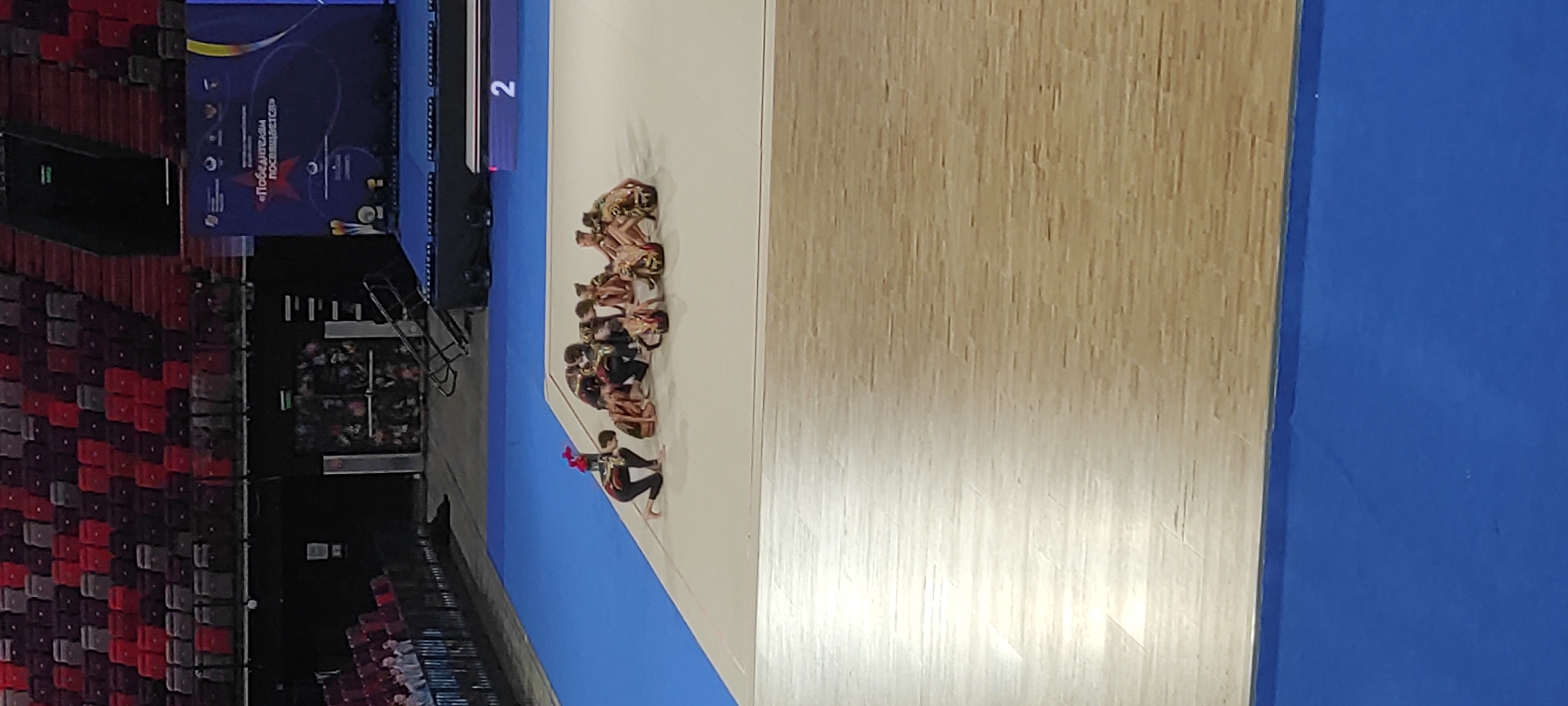 The image depicts a group of young female gymnasts in rhythmic gymnastics attire, gathered on a white mat with a blue border in what appears to be an indoor arena. One gymnast, kneeling on the left side of the mat, is holding a bouquet of red flowers. The gymnasts are in various poses, some sitting, some crouched, and they seem to be interacting with each other. In the background, there are rows of empty red and grey seats typical of a sports venue. A large blue banner with text and graphics, potentially indicating an event or sponsorship, is visible in the upper right background. A digital scoreboard displaying the number "2" is also present. The floor in the foreground is a polished wooden surface. The overall scene suggests a competition or performance event. The location is specified as Moscow, Russia, and based on the banner, the event is likely related to the "Festival of Gymnastics" as suggested by the Russian text "Festival of Gymnastics dedicated to the winners."