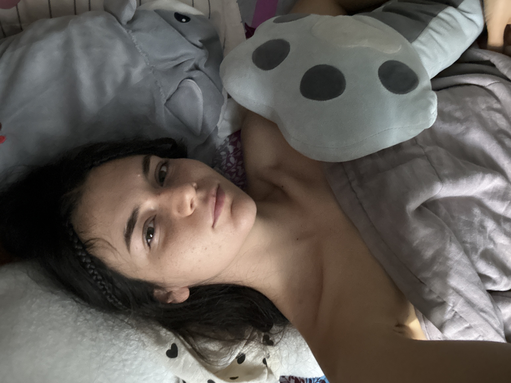 A young woman is captured in a close-up, high-angle shot, lying in a bed, likely in a bedroom in Mũi Né, Vietnam. She is looking directly at the camera with a neutral to slightly contemplative expression. Her dark hair, which includes some braided strands, frames her face, revealing natural freckles. Her upper body is partially visible, seemingly without clothing, suggesting she is under the covers or wearing minimal sleepwear.
She is surrounded by plush toys and bedding. To her left (viewer's right in the image), a large grey plush toy, possibly a shark or similar creature with small black eyes, rests next to her head. Above her right shoulder (viewer's left), another light grey plush toy, shaped like a mushroom with distinct dark grey circular spots, is visible.
The bedding includes a white pillow adorned with small black heart patterns beneath her head. A soft, light grey blanket covers part of her body and arm, while a patterned reddish-purple fabric is also visible near her shoulder. The overall lighting is soft and diffuse, consistent with an indoor daytime setting, possibly morning. The scene conveys a moment of rest or waking. There is no visible text within the image.