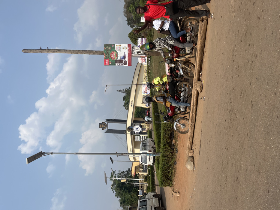 This image captures an outdoor street scene under clear daylight, depicting the entrance to the University of Ilorin in Odan Asun, Nigeria.
The foreground consists of a dark asphalt road surface on the left and a dirt shoulder on the right, featuring two rough-hewn wooden logs laid horizontally and several rocks.
In the mid-ground, five individuals are present near three motorcycles. On the right, a male in a red short-sleeved polo shirt, dark trousers, and a black shoulder bag stands, facing left with his right arm raised and hand open in a gesture. To his left, a red motorcycle is positioned with two occupants: one individual in a green helmet and a black and white patterned long-sleeved top seated at the front, and a female in a white short-sleeved top and blue jeans seated at the rear. Further left, two additional motorcycles are present. One is red, occupied by a male in a dark helmet, a yellow high-visibility vest over a dark shirt, and blue jeans. Adjacent to it, a lighter-colored motorcycle is occupied by another male in a dark helmet, a yellow high-visibility vest over a dark shirt, and blue jeans.
Behind the motorcycles and the dirt shoulder, a manicured green hedge forms a barrier. Several modern streetlights with solar panel tops are visible. A tall, bare wooden utility pole stands on the right, displaying a large square advertising banner with red and green festive graphics, text including "Wole Mobolaji" and "Merry Christmas," and images of electronic products.
The background prominently features the main entrance to the University of Ilorin. This entrance consists of a curving, light-colored gate structure with "UNILORIN" branding visible on multiple signs. Centered above the gate is a tall, dark, arch-like structure surmounted by a university crest or emblem. Beyond the gate, two white vans or minibuses are visible on a roadway, and additional greenery and distant buildings are present.
The sky is light blue with scattered white cumulus clouds.