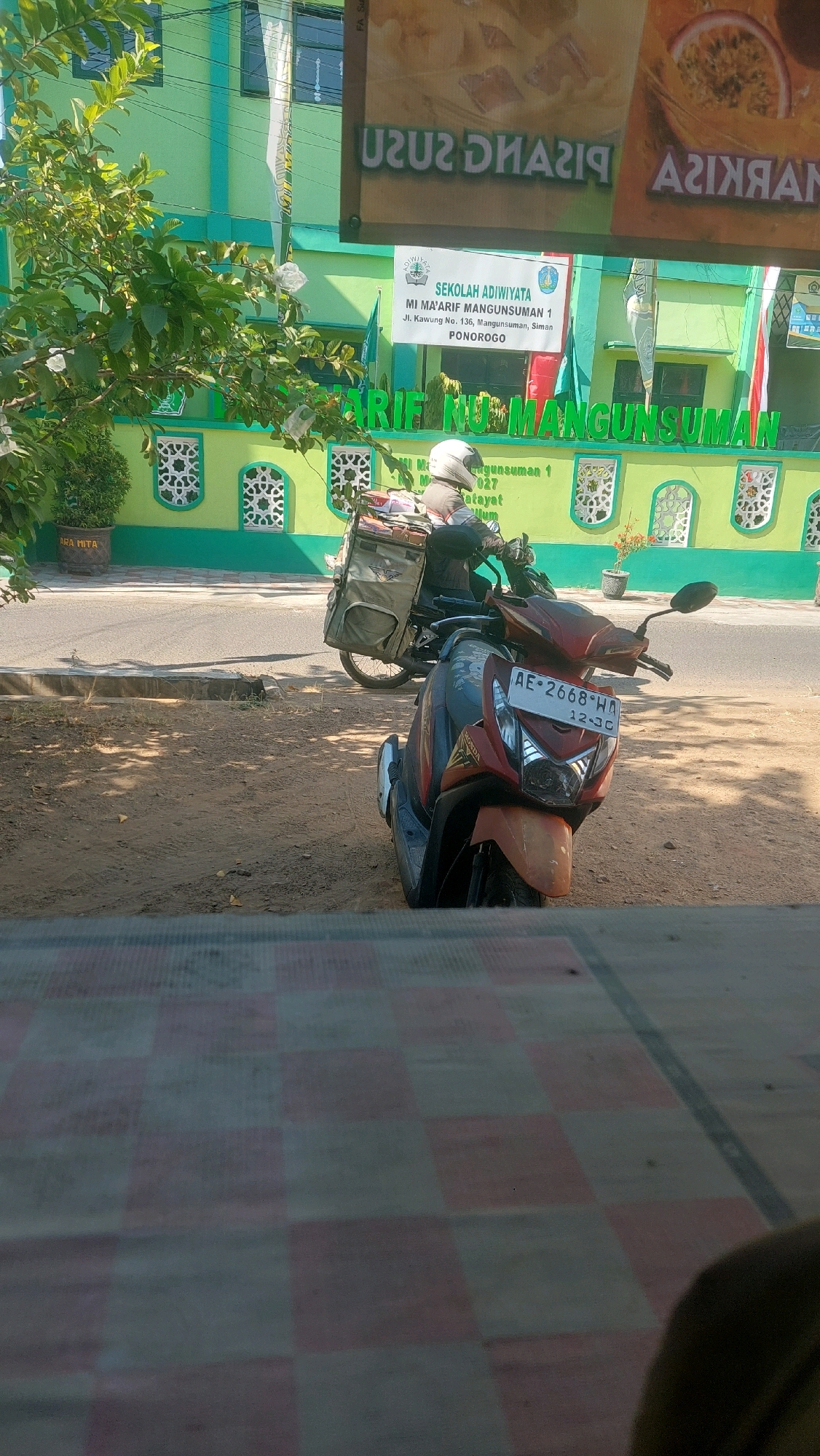 In the city of Mangunsuman, Indonesia, a motorcycle with license plate AE 2668 WA is parked on a dirt area in front of a light green building. The building has signage indicating it is a school, "SEKOLAH ADIWİYATA MI MA'ARIF MANGUNSUMAN 1" and mentions "Jl. Kawung No. 136, Mangunsuman, Simo, Pondorogo." The school building has decorative white windows. A person wearing a white helmet is seated on another motorcycle parked further back, a large, olive-green cargo carrier attached to its rear. The foreground of the image shows a checkered pattern, possibly a mat or tablecloth. Sunlight casts shadows on the ground, indicating a bright day. A tree with green leaves is visible on the left side of the image.