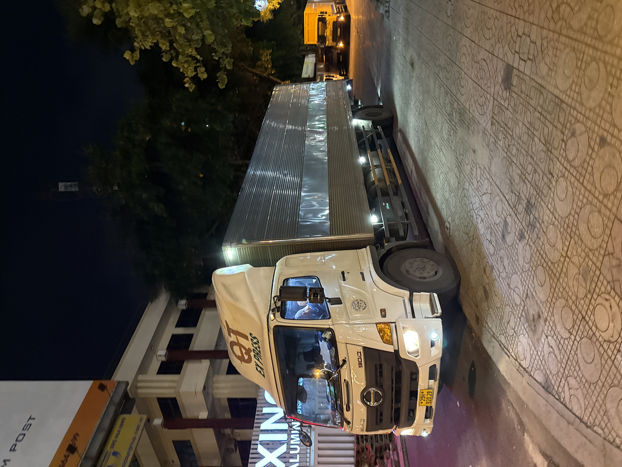 A white Hino truck with "QT EXPRESS" branding is parked on a sidewalk next to a street at night in Nha Trang, Vietnam. The truck has a large silver cargo box and its headlights are illuminated. A yellow truck with "NGUYEN VIET NAM" written on its side is visible parked behind the white truck, further down the street. The sidewalk is paved with patterned tiles, and there are trees and buildings in the background. A portion of a large white billboard with black text is visible in the upper left corner of the image. The scene is illuminated by streetlights and the truck's headlights.