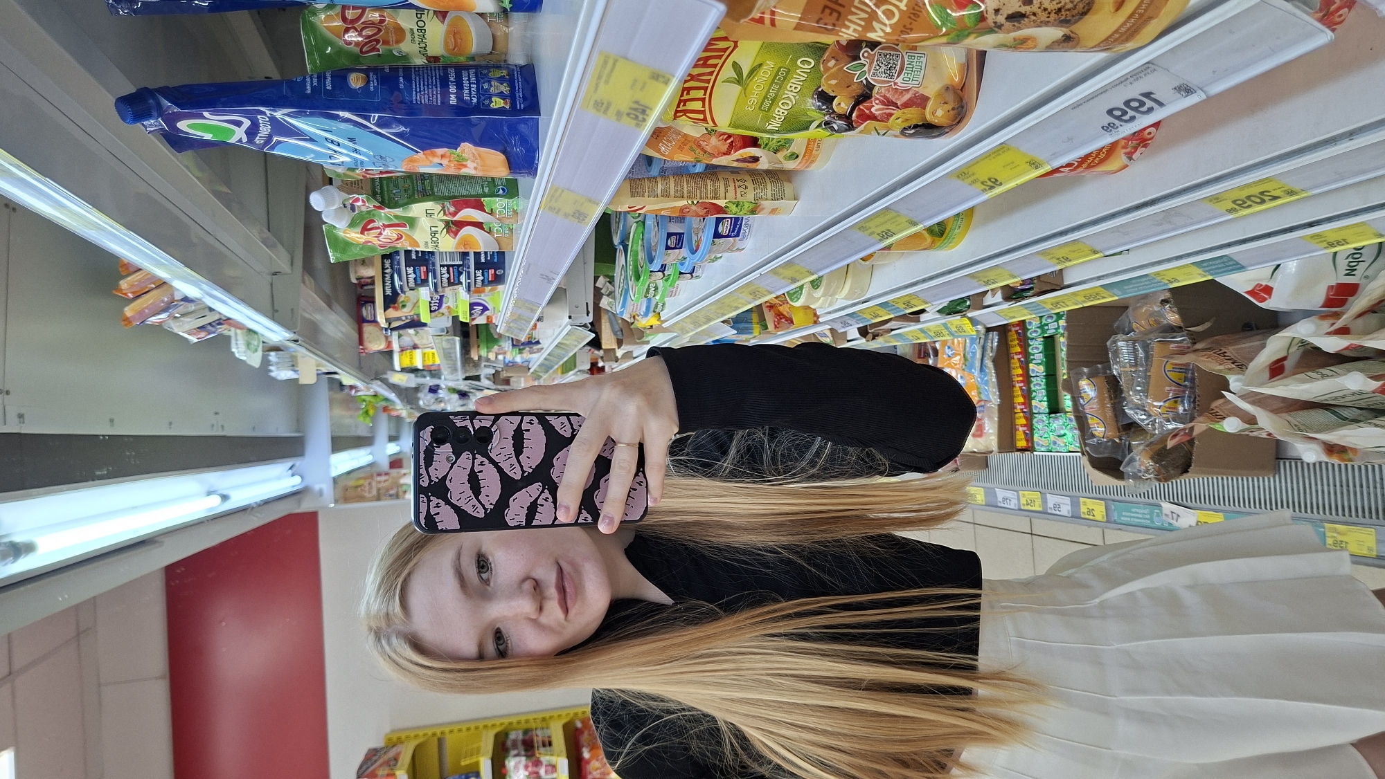 A young woman with long blonde hair and light skin, wearing a black long-sleeved shirt and a white pleated skirt, stands in a grocery store aisle. She is holding a smartphone in her right hand, which is positioned to take a selfie. The phone case features a pattern of pink lips. She has a light dusting of freckles across her nose and cheeks. Her expression is neutral. The background consists of shelves stocked with various food products, including packaged snacks, sauces, and dairy items. Fluorescent lights illuminate the scene from above. The overall environment suggests a brightly lit retail space. The location is identified as a supermarket in Chaikovsky, Russia.