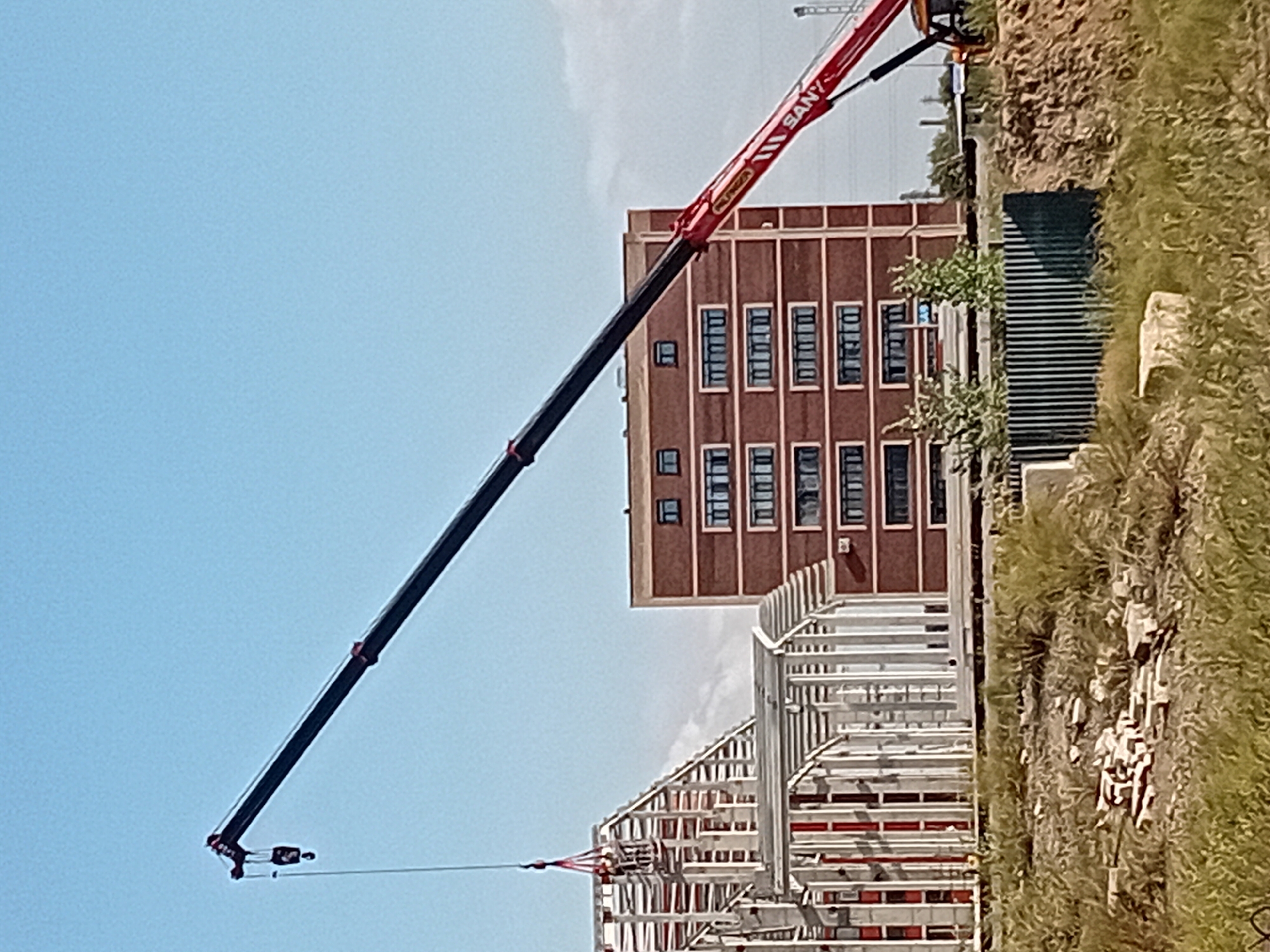 The image depicts a construction site under a clear blue sky in Semey, Kazakhstan. A large red and black crane extends diagonally across the frame, its boom reaching towards the upper left. The crane's hook is visible, with a load being lifted towards a partially constructed building on the left. This building features a skeletal framework of white beams and red vertical supports. To the right of the construction, a multi-story brick building with rows of dark windows stands completed. The foreground is occupied by a dirt embankment covered with dry grass and scattered debris, including what appear to be bricks. A dark green metal fence runs along the middle ground behind the embankment. The overall scene suggests ongoing construction activity within an urban or semi-urban environment.