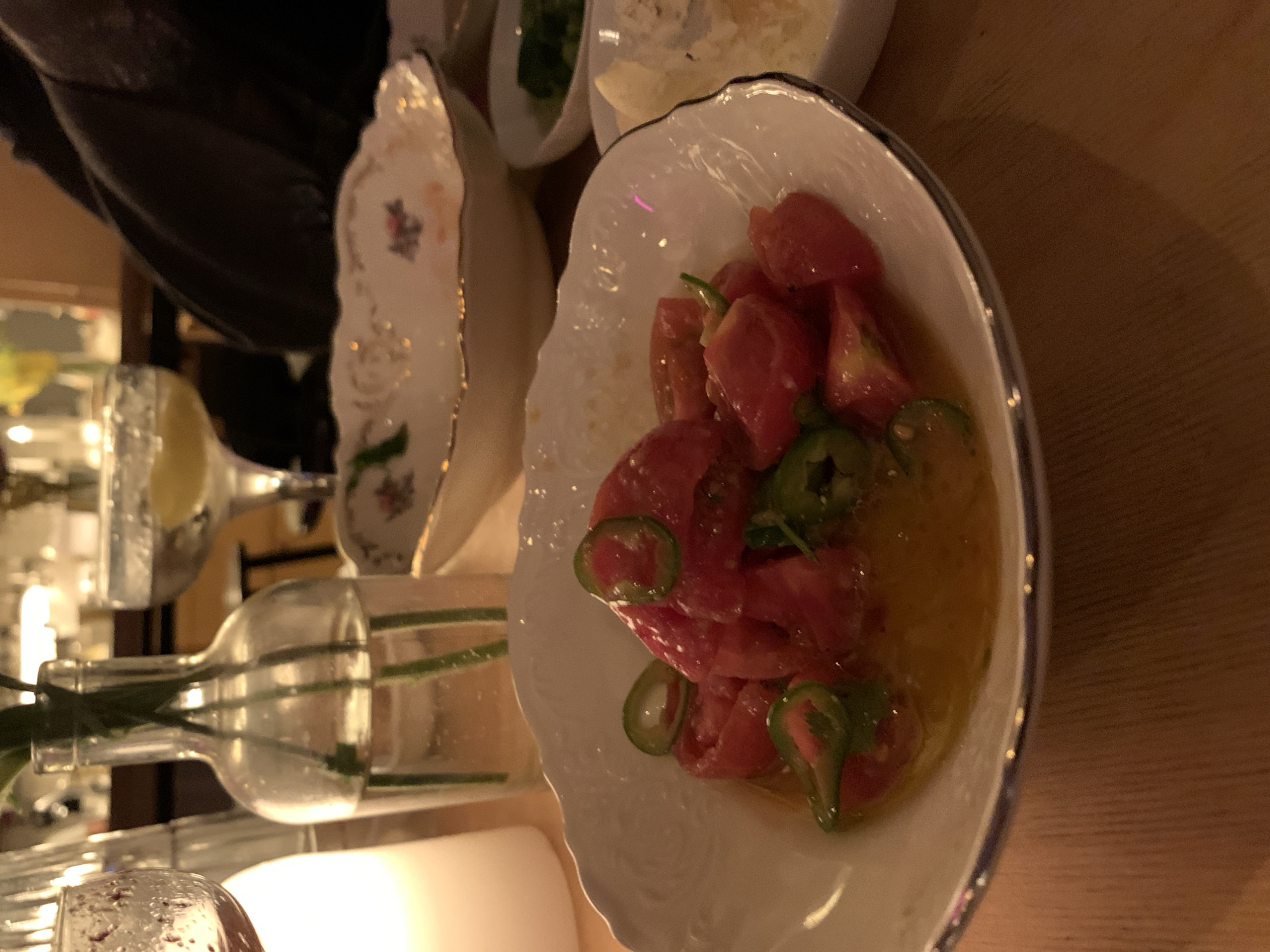 This image captures a close-up view of a dining table in a dimly lit indoor setting, likely a restaurant or bar in Tel-Aviv, Israel. The central focus is a decorative white bowl with scalloped, gold-rimmed edges, holding a vibrant dish composed of diced red tomatoes and green chili slices immersed in a yellowish-orange liquid, possibly a dressing or sauce.
To the left of this main dish, another similar white ornate plate or bowl is partially visible. In the background, two small white bowls contain what appears to be grated cheese and chopped green herbs. A clear glass bottle holding green plant stems and a cocktail glass with a yellow drink and a lemon slice are also present, indicating beverages are being served. A white cylindrical object, likely a candle or a small lamp, casts a warm glow on the wooden table surface, contributing to a cozy, evening atmosphere. A dark-clothed figure is partially visible in the upper left corner, suggesting someone is seated at the table. The scene depicts a relaxed dining or social event taking place at night. There is no visible text in the image.
