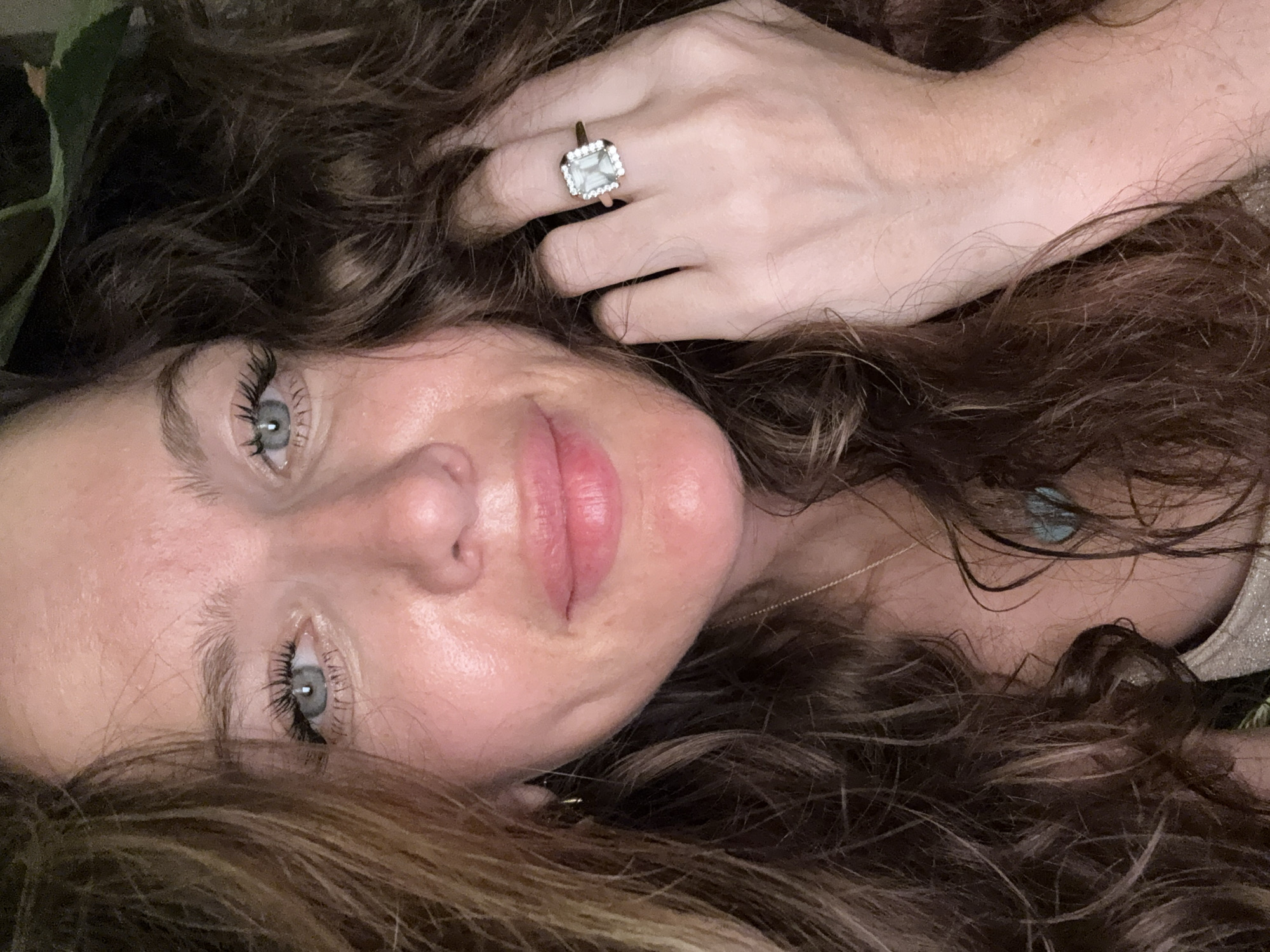 A close-up, chest-up portrait of a woman with curly brown hair is featured against a dark, out-of-focus background. She has fair skin, blue eyes, and is wearing a silver top. Her right hand, adorned with a ring featuring a square-cut stone surrounded by smaller stones, is raised near her face, with her fingers partially obscuring her hair. The image does not provide any information about the location in Dubai, United Arab Emirates.