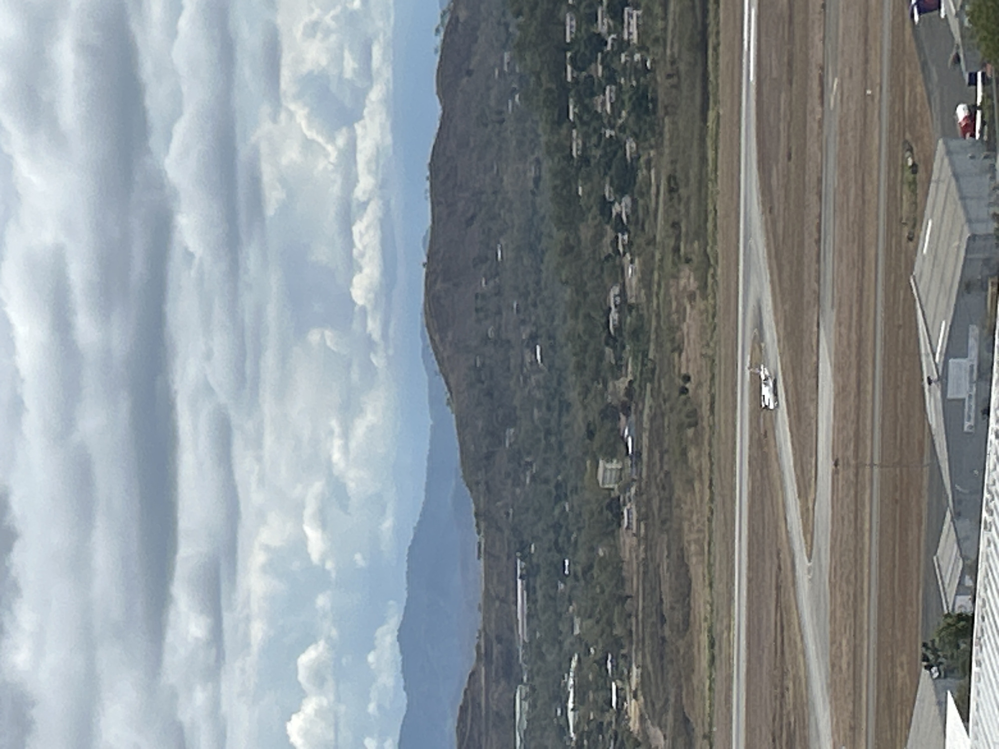 The image captures a view of an airport runway with a small airplane in motion, likely taxiing or preparing for takeoff. In the background, rolling hills covered in vegetation rise, dotted with scattered buildings, suggesting a semi-urban or suburban environment. A hazy sky with scattered clouds looms above the landscape. The foreground shows the edge of airport infrastructure, including buildings with corrugated roofs and some vehicles. The scene appears to be set in a warm, possibly tropical climate, given the lush greenery and the general atmosphere. The lighting suggests it is daytime, with diffused sunlight filtering through the clouds. The overall impression is one of a functional airport in a scenic, somewhat undeveloped, or developing area.