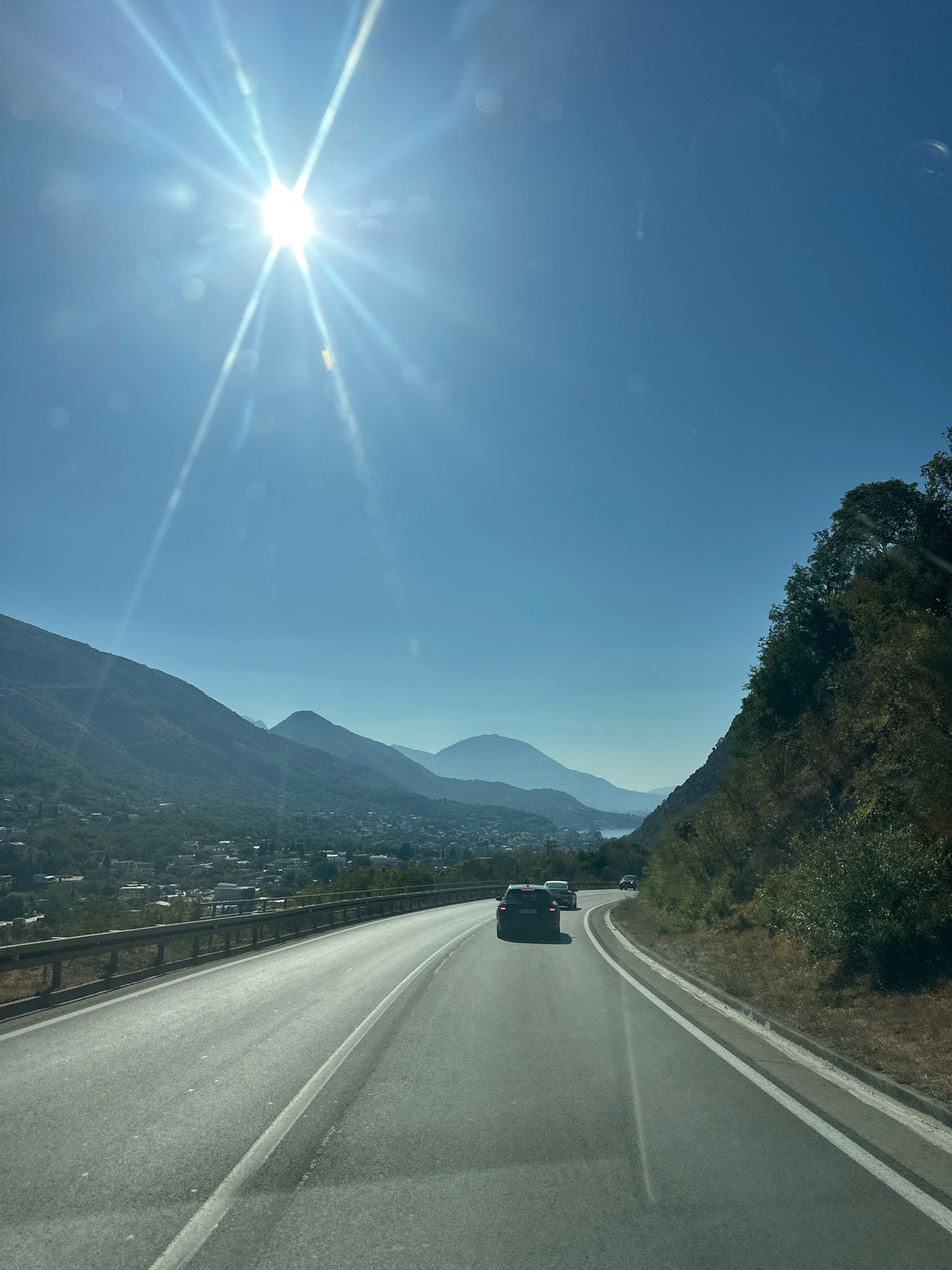 The image shows a view from a vehicle driving on a road that curves to the right, overlooking a town nestled in a valley below mountains. The sun is bright and high in a clear blue sky, casting lens flares across the upper portion of the image. The road has two lanes, with a guardrail on the left side separating it from a steep embankment. Several vehicles are visible on the road ahead, including a black SUV in the left lane and at least two other cars in the right lane.
The town below appears to be Zagrađe, Montenegro, characterized by numerous buildings with red-tiled roofs spread across the lower slopes of the mountains. The mountains themselves are covered in lush green vegetation and rise dramatically in the background. In the distance, hazy outlines of more mountains and possibly a body of water can be seen. The right side of the road is bordered by a densely vegetated hillside with trees and shrubs. The overall scene depicts a scenic drive through a mountainous coastal region.