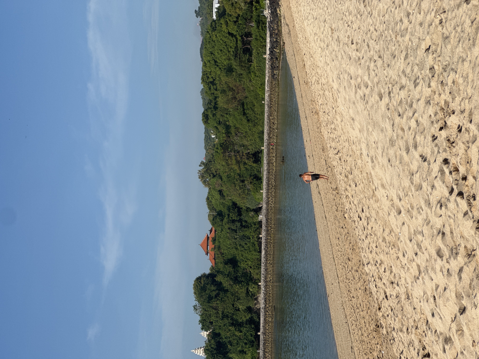 The image captures a coastal scene in Unknown, Indonesia. A sandy beach occupies the foreground, marked with footprints. A seawall separates the beach from the water. A lone individual stands on the beach, close to the water's edge. Behind the seawall, dense green vegetation covers the land. The tops of buildings, one with an orange roof, are visible amongst the trees. The sky above is a clear blue with scattered clouds.