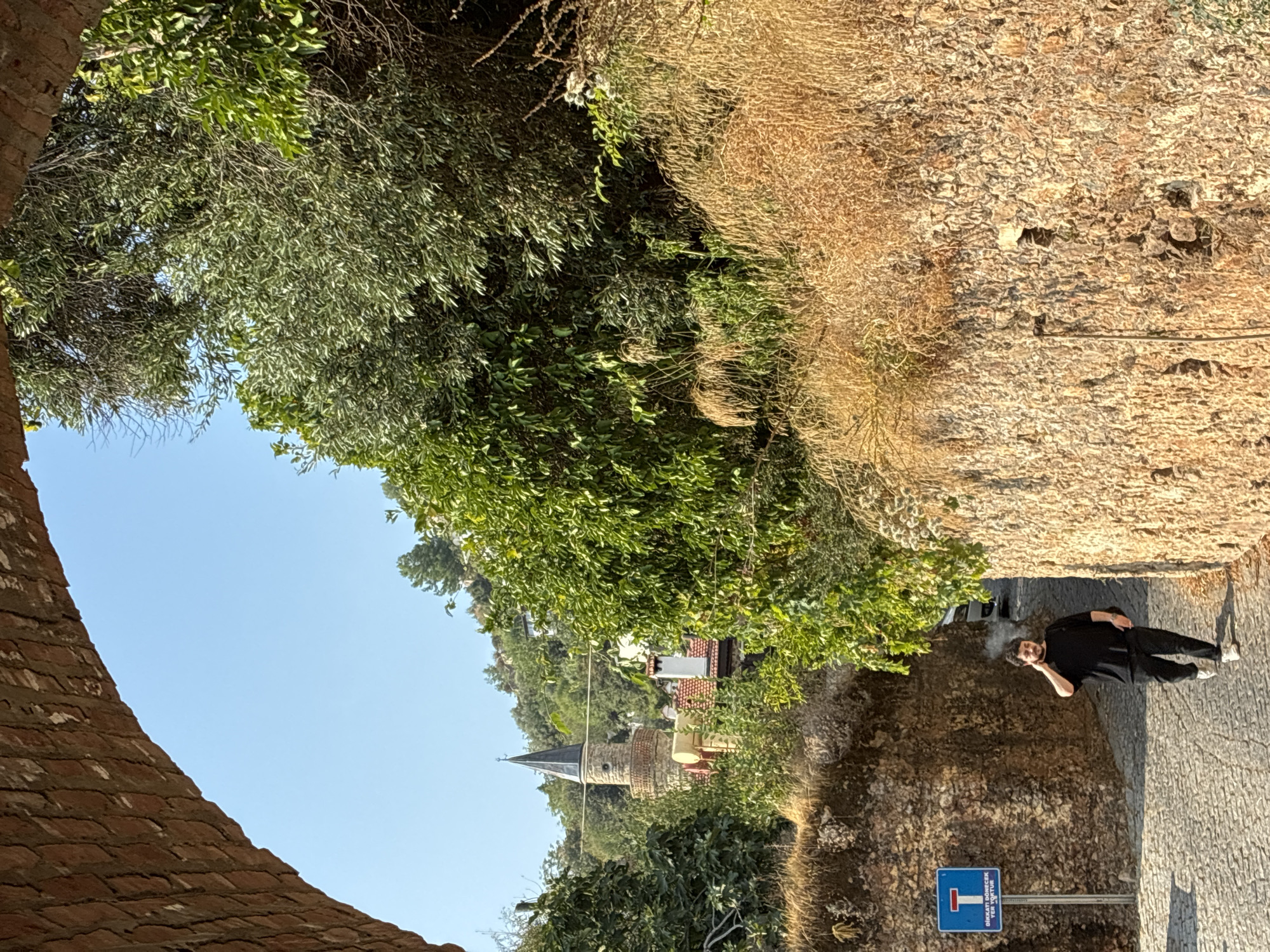 The image depicts a narrow, cobblestone street in Alanya, Turkey, framed by an arched brick structure on the left and a rough stone wall on the right. Lush green foliage and dry, tall grass line the upper portions of the walls, with a minaret visible in the distance amidst the trees and hillside homes. A young man wearing a black t-shirt and black pants walks towards the camera on the street, holding a cell phone to his ear and with what appears to be smoke emanating from his mouth. A blue traffic sign with a red "T" symbol and Turkish text is posted on the left side of the street. Sunlight casts shadows on the cobblestones, indicating a bright, sunny day.