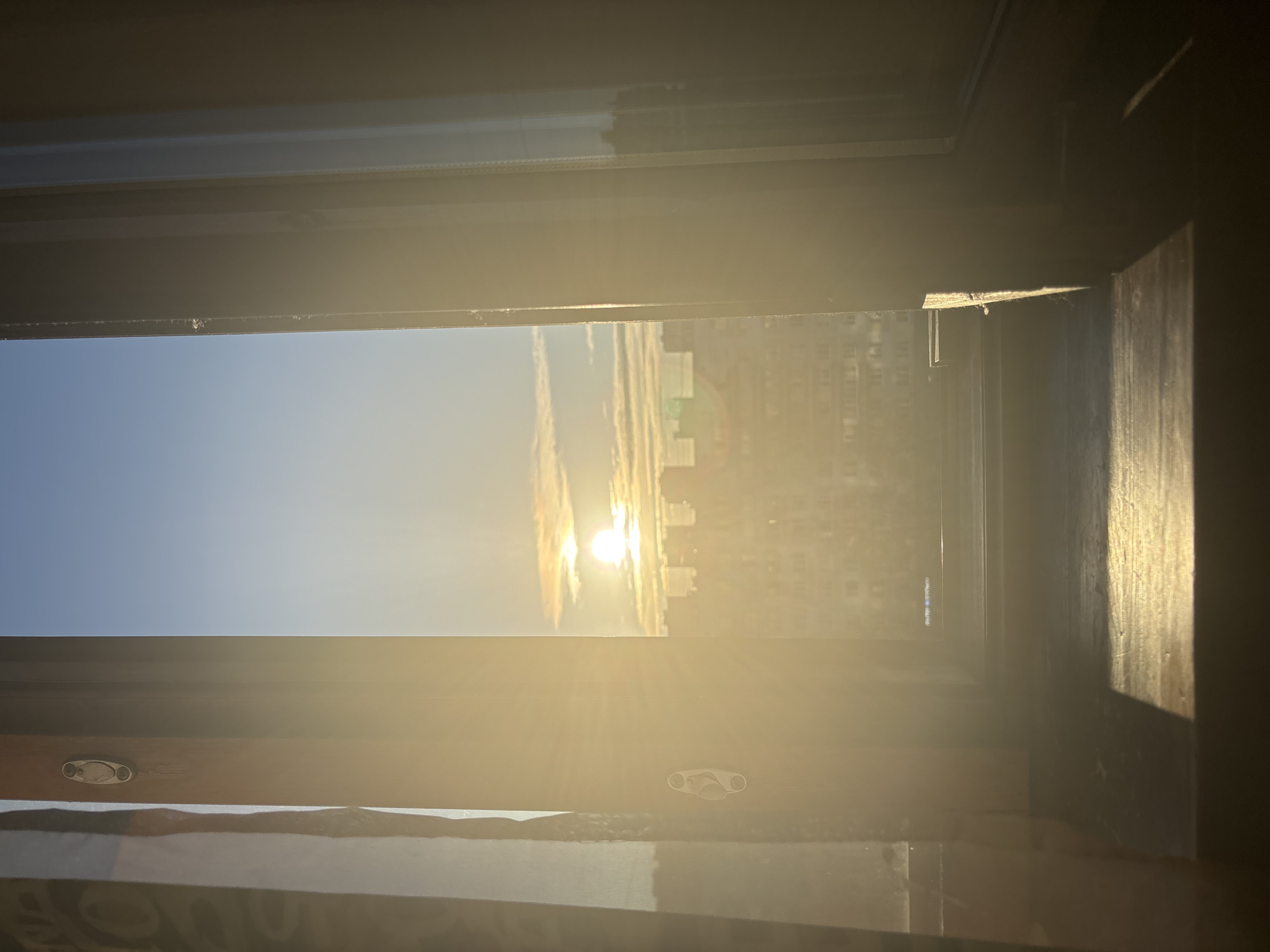 The image shows a sunrise or sunset seen through a window. The sky is blue with some light clouds around the sun. Buildings are visible in the distance, obscured by the bright light. The window frame is partially visible, and some curtains are at the bottom of the frame. The location is inferred to be in the city of Unknown, Russia.
