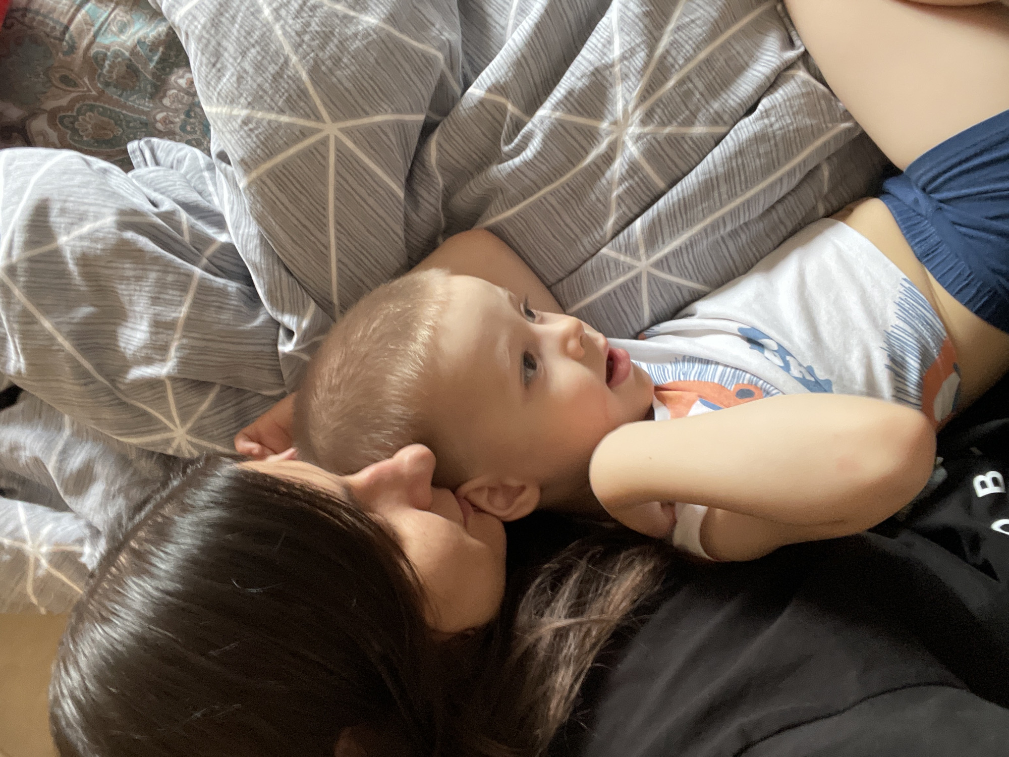 An intimate, top-down shot captures an adult and a young child lying close together on a bed. The adult, with long dark brown hair, is seen from the side, with her face close to the child's head, possibly in an affectionate gesture like a kiss or a sniff. The child, who has light blonde hair, is lying on their back, looking upwards and to the side with an open mouth, appearing curious or engaged.
The child is wearing a white t-shirt with faint orange and blue patterns, and blue shorts. The adult is wearing a black top or t-shirt. The bedding consists of a patterned duvet or blanket with a gray and white geometric design, and another section of bedding featuring a darker, more elaborate paisley-like pattern in shades of blue, green, and brown.
The scene conveys a sense of warmth, closeness, and tender affection between the two individuals. A letter "B" and other partially visible letters are discernible on the adult's black shirt. The lighting suggests it is daytime, though no windows or specific location cues are visible.