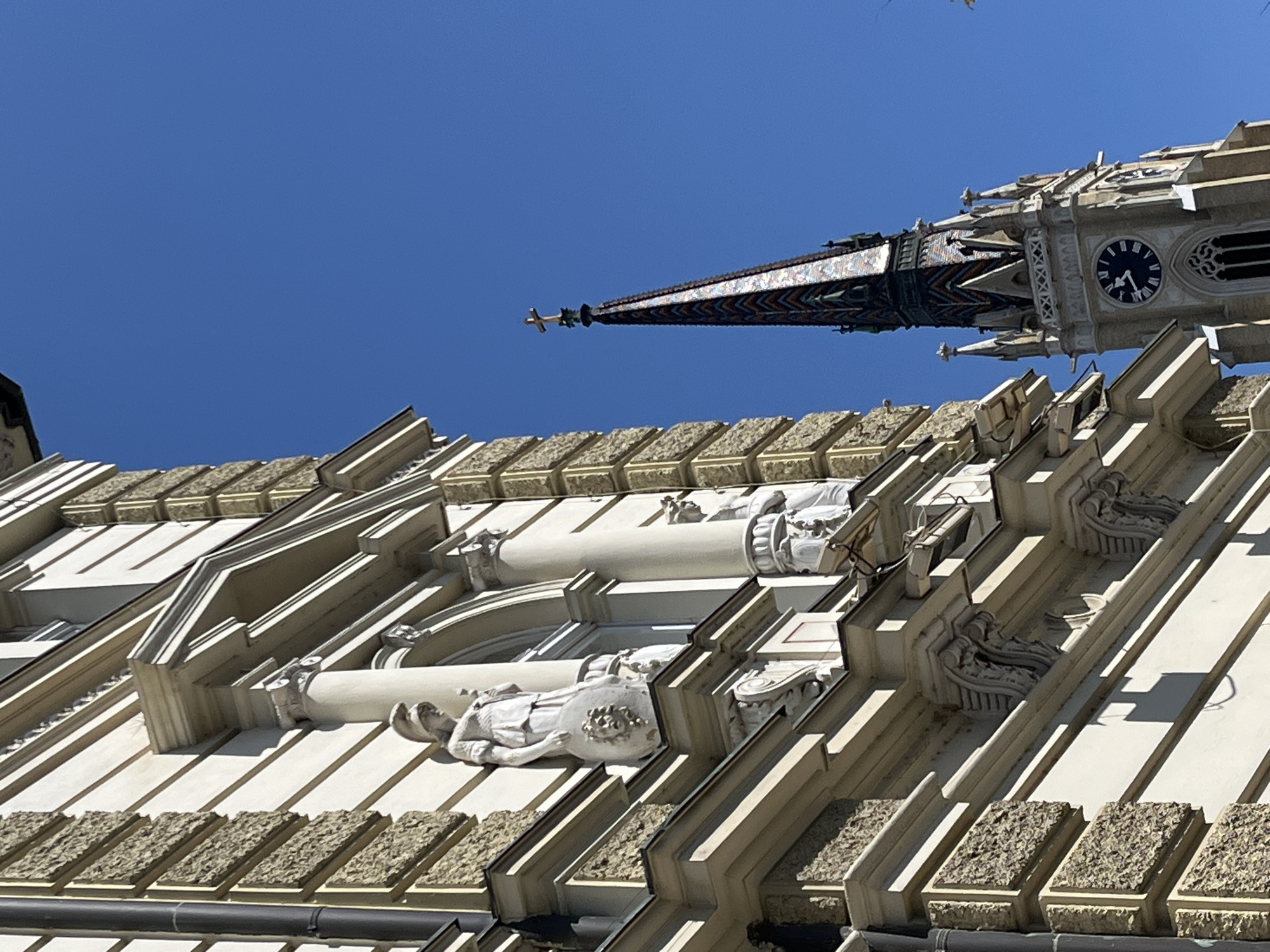 The image shows a low-angle view of architectural details of a building in Novi Sad, Serbia. The building features white stone or plaster facade with decorative elements. The architecture includes a clock tower with a black clock face and a decorative spire tiled in various colors, with a gold cross on top. Below the tower, decorative pillars and sculptures are seen, with the building's roof visible in the foreground. The sky is clear and blue.