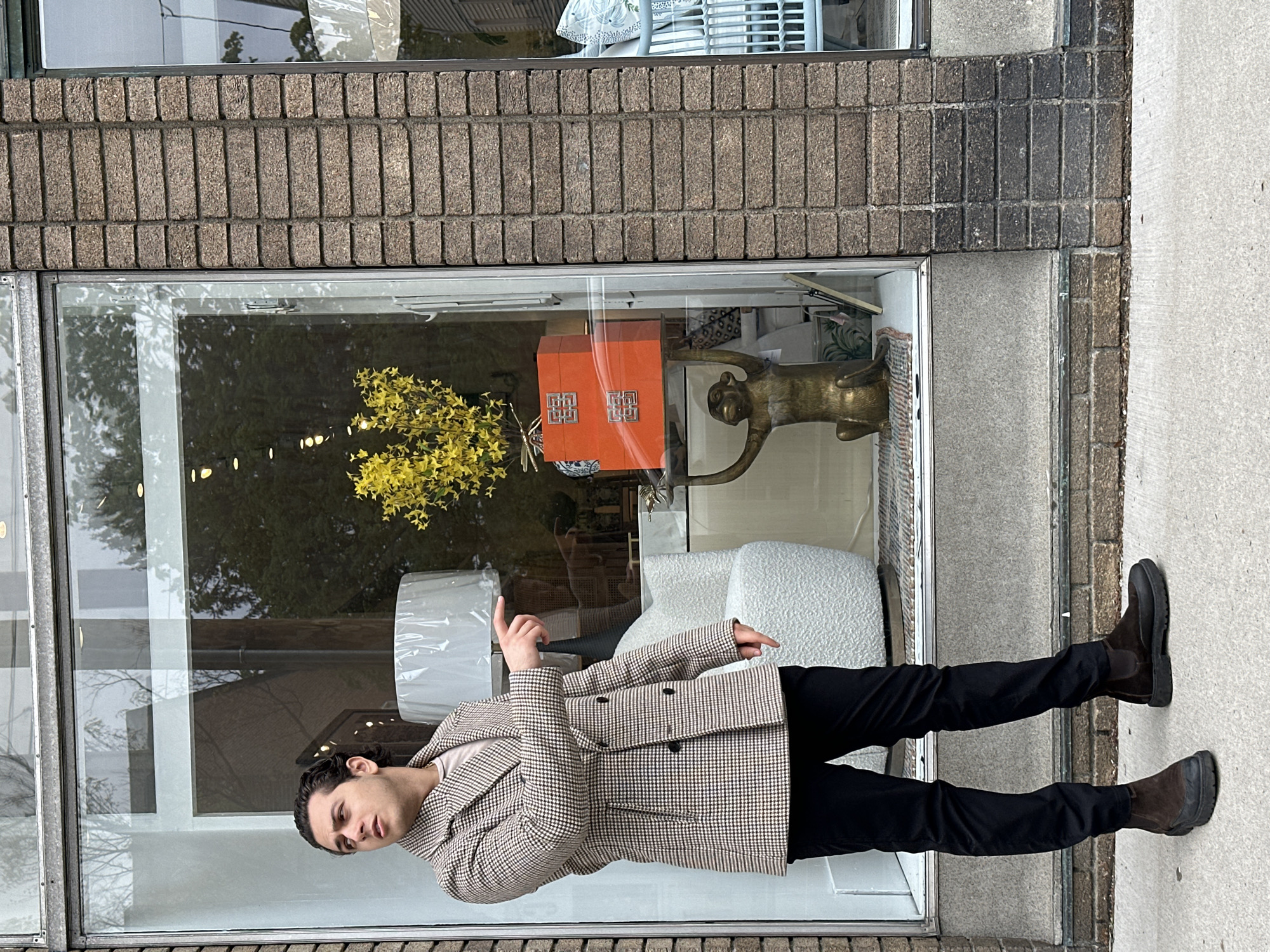 A young man stands outside a shop window in the Village of Cold Spring, United States of America, during what appears to be an overcast day. He has dark, shoulder-length hair and is wearing a brown and white checked jacket over a light top, black trousers, and dark brown Chelsea boots. He is pointing with his right hand into the shop window, looking towards his left with a neutral expression.
The shop window displays several interior decor items. To the left, a large white drum lampshade is visible, next to a textured white armchair or small sofa. A vibrant yellow floral arrangement, possibly forsythia, adds a splash of color. Central to the display is an orange box or cabinet featuring prominent silver-colored, intricate geometric designs resembling interlocking letters on its two visible doors. To the right, a striking bronze-colored statue of a monkey with an extended arm adds an artistic touch.
The building's facade above and surrounding the window is constructed from rectangular, rough-textured, dark grey-brown bricks laid in vertical stacks. The man stands on a light grey concrete sidewalk. Reflections in the window reveal trees and the muted light of an overcast sky. No discernible text is clearly visible other than the stylized emblems on the orange box.