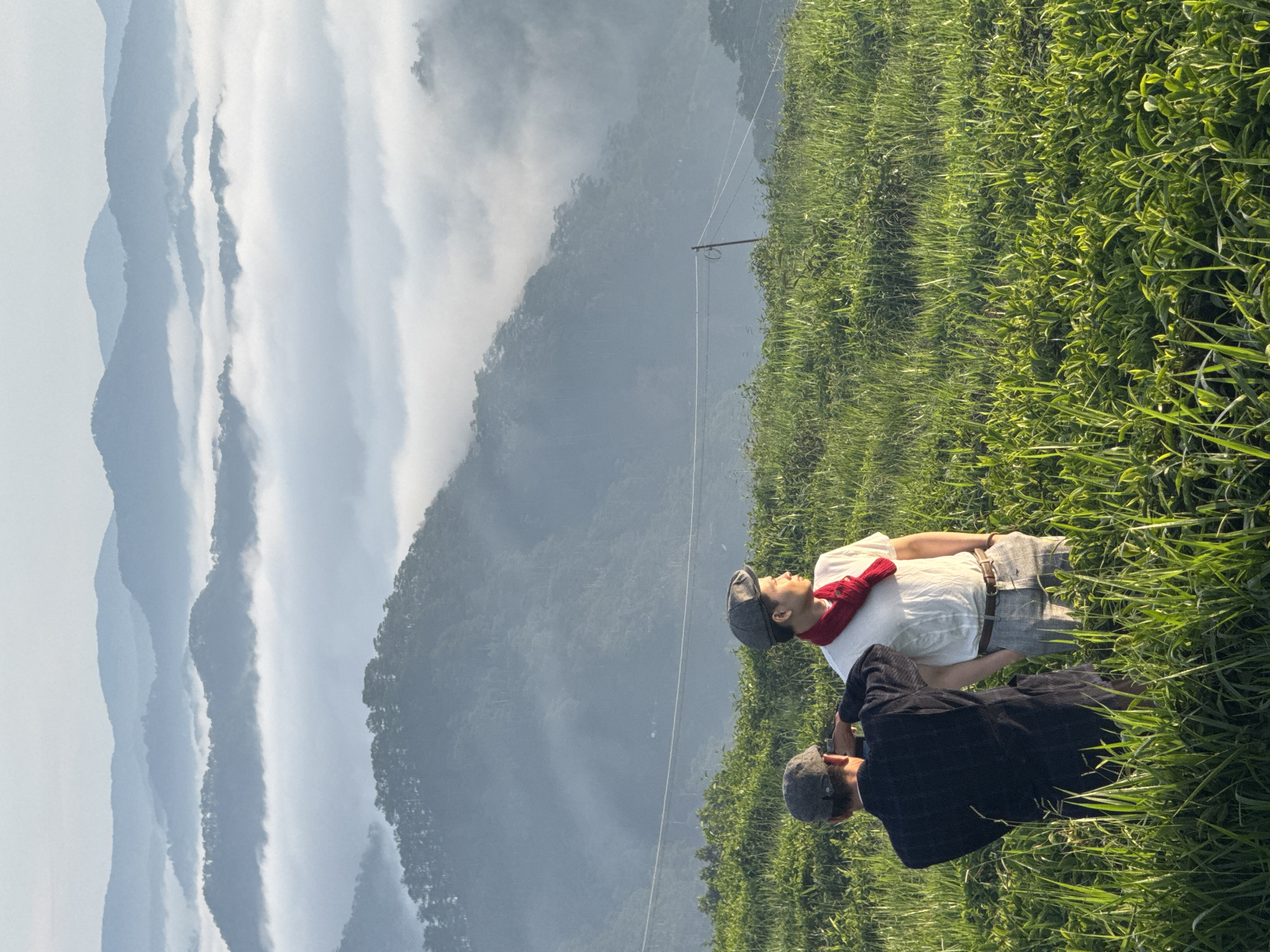Two individuals stand in a grassy field, facing away from the camera and looking out at a misty mountain landscape. The person on the left wears a dark jacket and a grey flat cap. The person on the right wears a white t-shirt, a red scarf, grey pants, and a grey flat cap. They are positioned on a hillside covered in lush green grass and some lower-lying bushes. In the background, multiple layers of mountains recede into the distance, partially obscured by a thick layer of white mist that creates a serene, ethereal atmosphere. The sky above is bright and hazy, suggesting early morning light. The overall scene evokes a sense of tranquility and contemplation, with the mist softening the hard lines of the mountains and creating a dreamy backdrop for the two figures. The image captures a moment of quiet observation of the natural beauty of the landscape. The location is identified as Phát Chi, Vietnam.