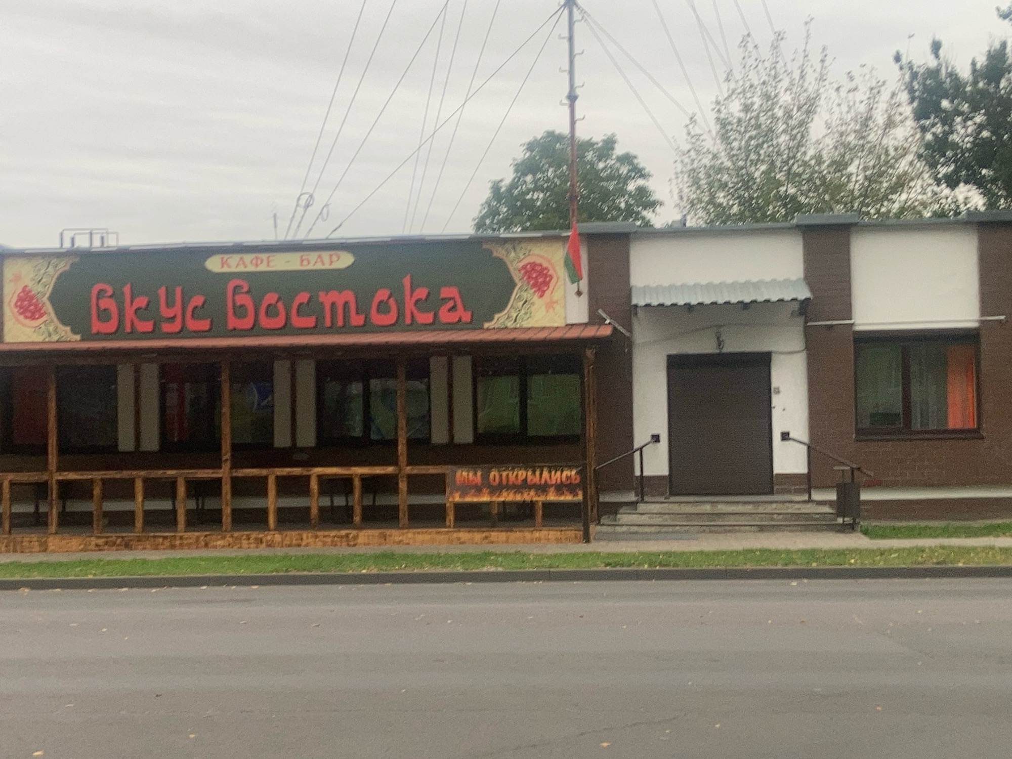 The image shows a cafe-bar named "Вкус Востока" (Taste of the East) located in Brest, Belarus. The establishment has a covered outdoor seating area with wooden railings and tables visible behind them. A banner above the entrance reads "МЫ ОТКРЫЛИСЬ" (We are open). The building features a dark brown facade with white and brown accents, and several windows. A small flag, possibly of Tajikistan, is attached to a pole above the entrance. The scene is captured from across a paved road, with a sidewalk and grassy area in between. The sky is overcast.