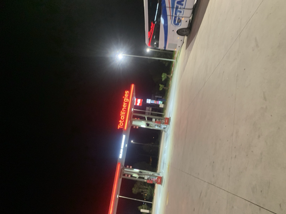 The image captures a TotalEnergies gas station at night in Khemisset, Morocco. The station's prominent canopy structure is brightly illuminated, displaying red "TotalEnergies" signage and white "SOLAR ENERGY" text. Below the canopy, multiple fuel pumps with red accents are visible, alongside a vertical price display board. The forecourt is a wide, light-colored paved surface, well-lit by overhead station lighting and multiple tall streetlights. A white and blue passenger bus, featuring "CTM" branding on its side and red accent lights, is stopped on the right side of the forecourt. The background shows a dark night sky and obscured vegetation. No individuals are present in the scene.