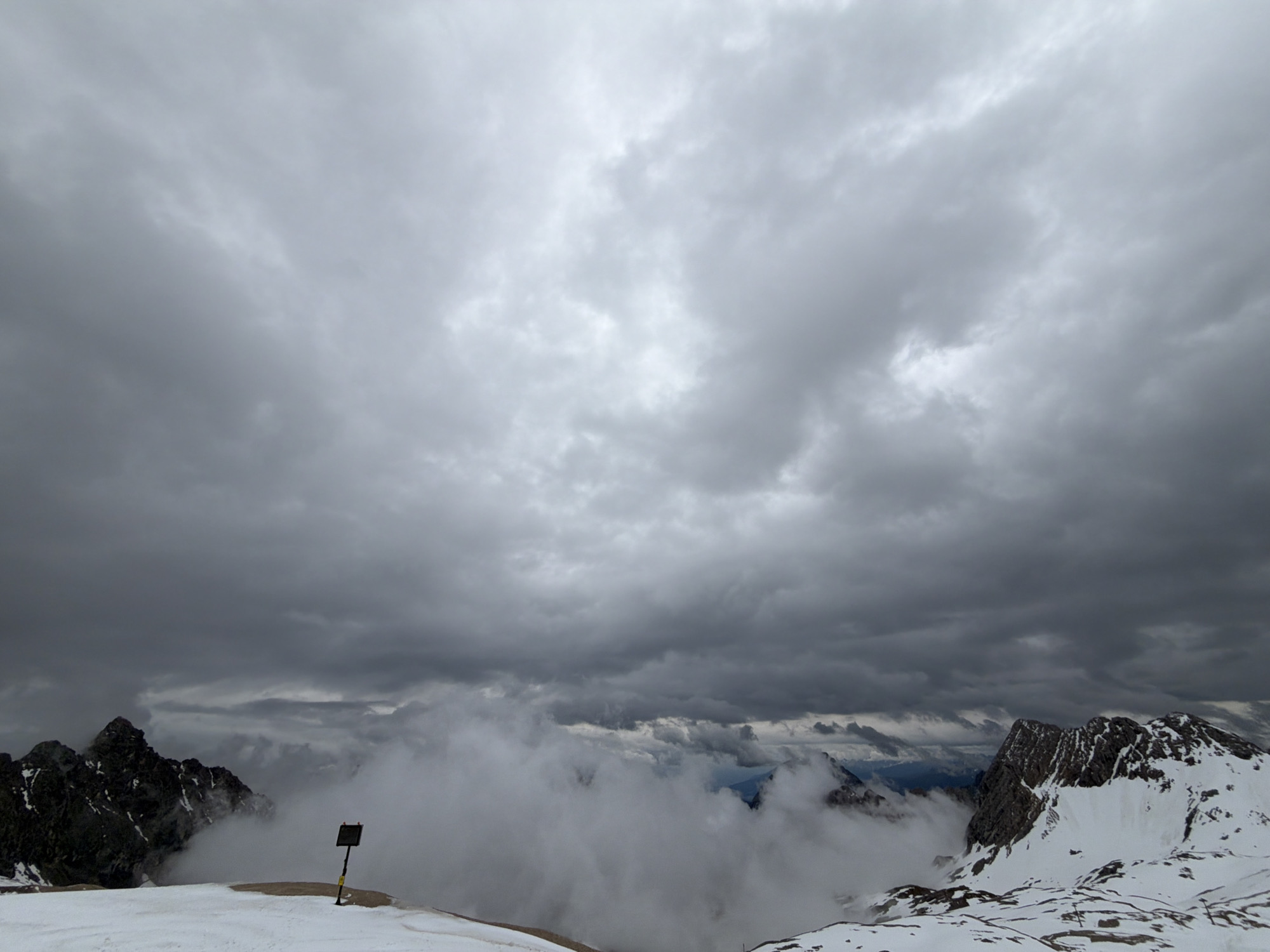 This image captures a high-altitude alpine landscape under a heavily overcast sky, likely in the vicinity of Garmisch-Partenkirchen, Germany, consistent with views from prominent peaks in the Bavarian Alps. The upper two-thirds of the frame are dominated by a dense canopy of dark grey stratocumulus clouds, exhibiting varied shades of grey with lighter patches indicating some thickness variations.
In the foreground, a snow-covered expanse with patches of bare, dark earth extends across the lower portion of the scene. Slightly to the left of the center, a dark, square-shaped sign is mounted on a yellow and black striped pole, planted firmly in the snow and earth. The sign's face is dark and provides no discernible text.
Immediately behind the foreground and to the left, rugged, dark rock peaks, partially dusted with snow, emerge from a dense layer of low-lying clouds or mist that obscures the middle ground. This cloud layer stretches across the central portion of the image, partially revealing additional snow-dusted, dark mountain formations and distant, hazy ridges towards the right side and in the background. No people or specific actions are visible within the frame. The overall environment conveys a cold, somber, and remote mountain setting.