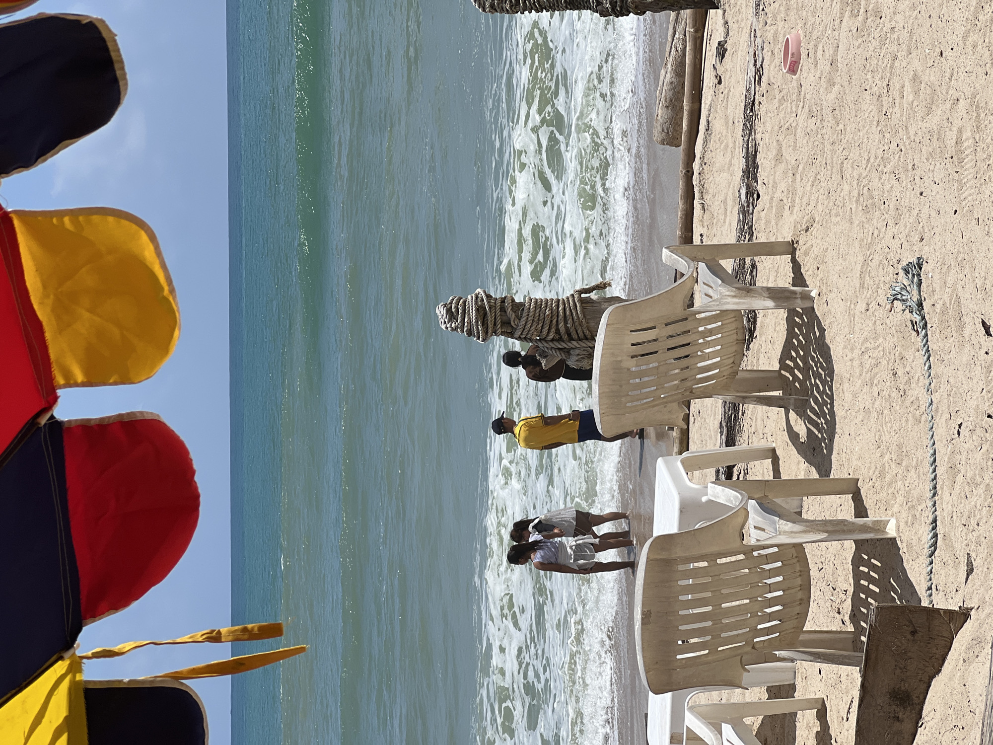 The image depicts a beach scene in Choeng Thale, Thailand. In the foreground, white plastic chairs are positioned on a sandy beach, with shadows cast by an overhead umbrella. In the mid-ground, several people are standing in the shallow water near the shore. Two young females, one in a striped top and white skirt and the other in a white top and brown skirt, are interacting. A male wearing a yellow shirt and blue shorts is also present, along with another individual partially obscured. The ocean exhibits moderate waves with white foam, extending to the horizon under a clear sky. The environment includes beach chairs, tables, a wooden post wrapped with rope, and scattered debris on the sand.