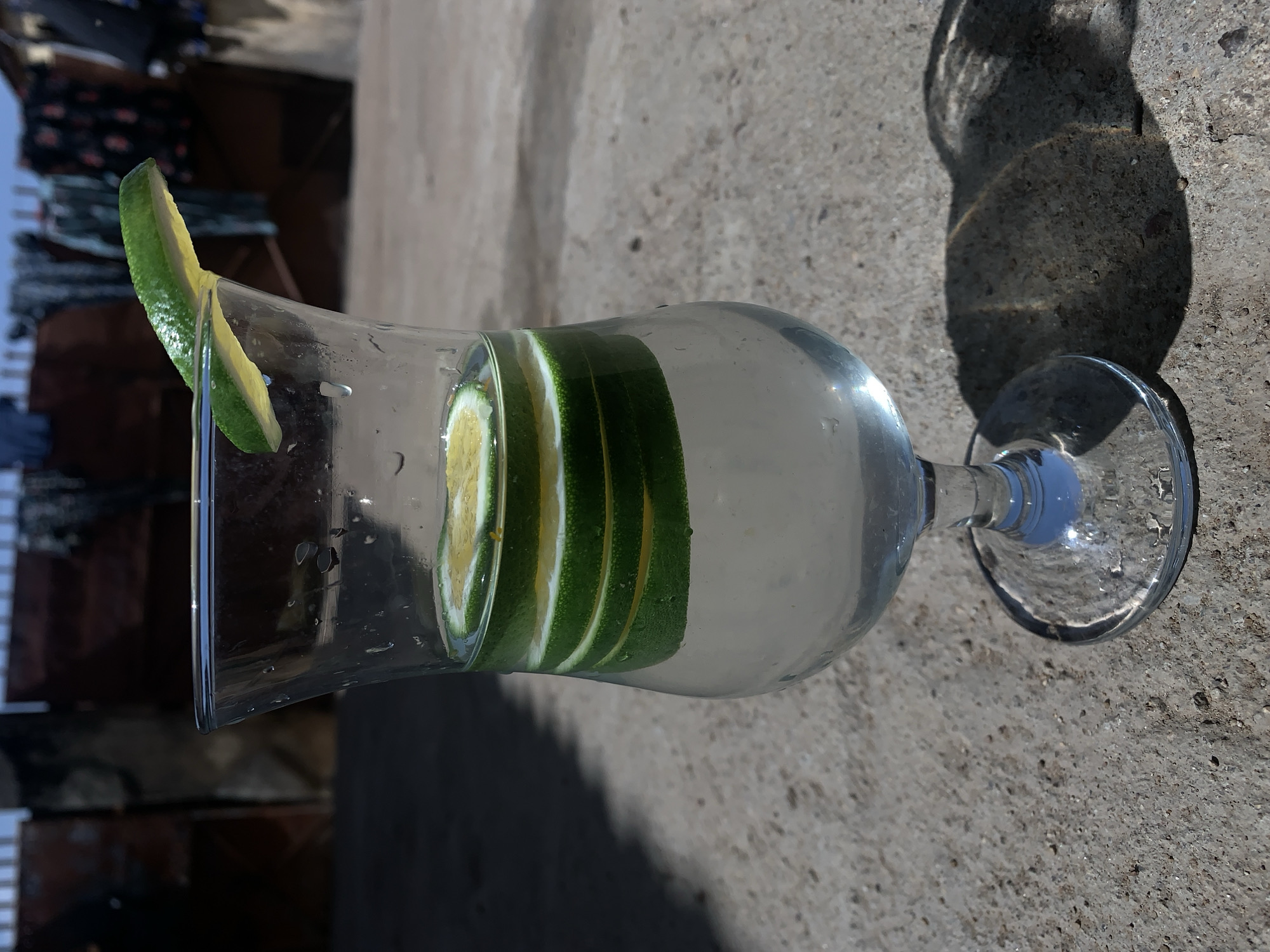 A glass of water with lime slices is on a concrete surface. A lime wedge garnishes the rim of the glass. The water in the glass is clear. In the background is a blurred area, with what appear to be dark objects or clothing hanging. The image was taken in Rafin Par, Nigeria.