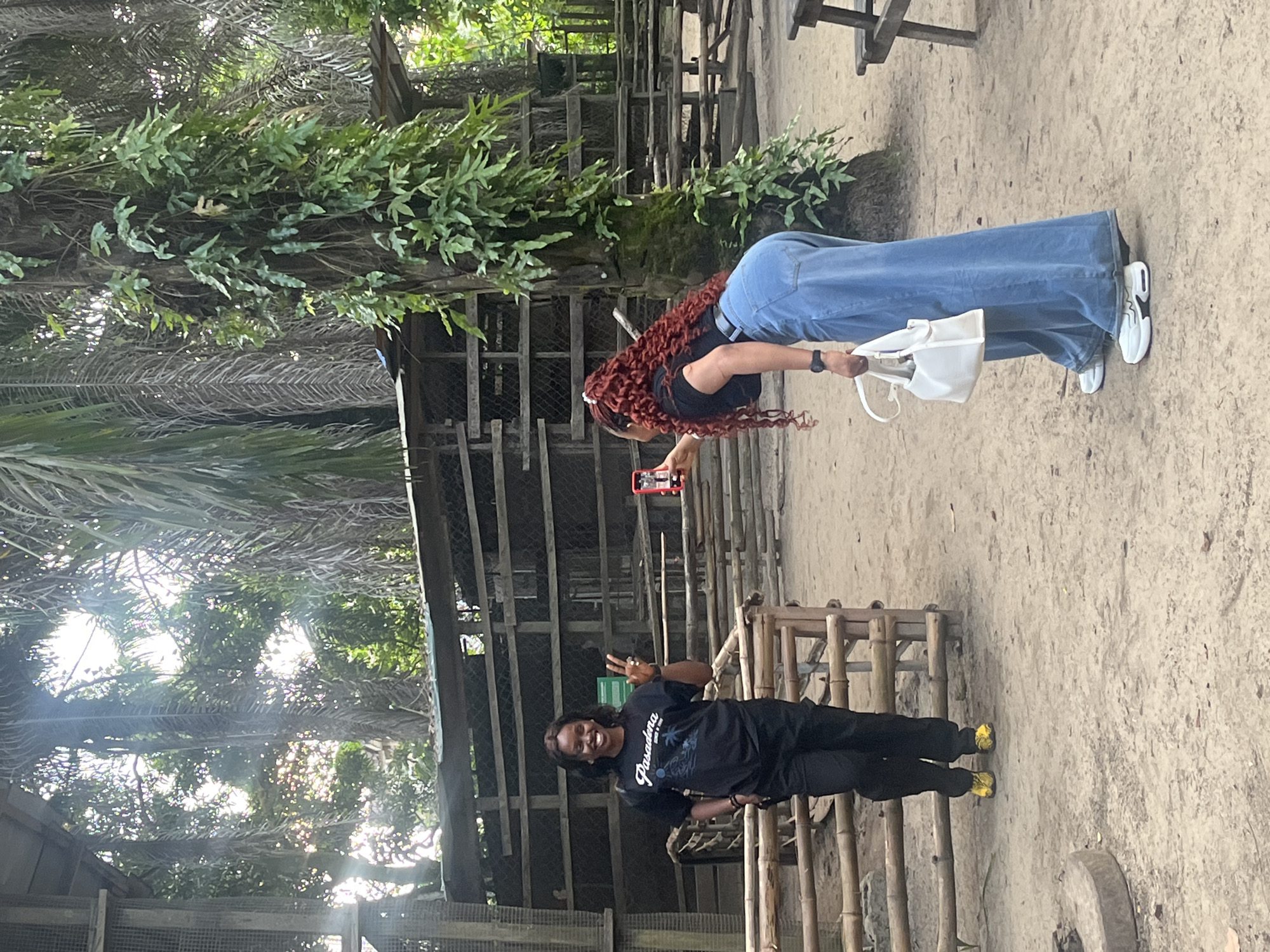 In Lekki, Nigeria, two women stand in an outdoor enclosure with a dirt ground and bamboo fences. A large palm tree with vines wraps around it is to the right. In the background, wooden structures with mesh and a picnic table are visible. The woman on the left, wearing a black t-shirt and black pants, smiles and holds up two fingers. The woman on the right, wearing a black top and wide-leg blue jeans, holds a red smartphone and takes a selfie. She also carries a white handbag.