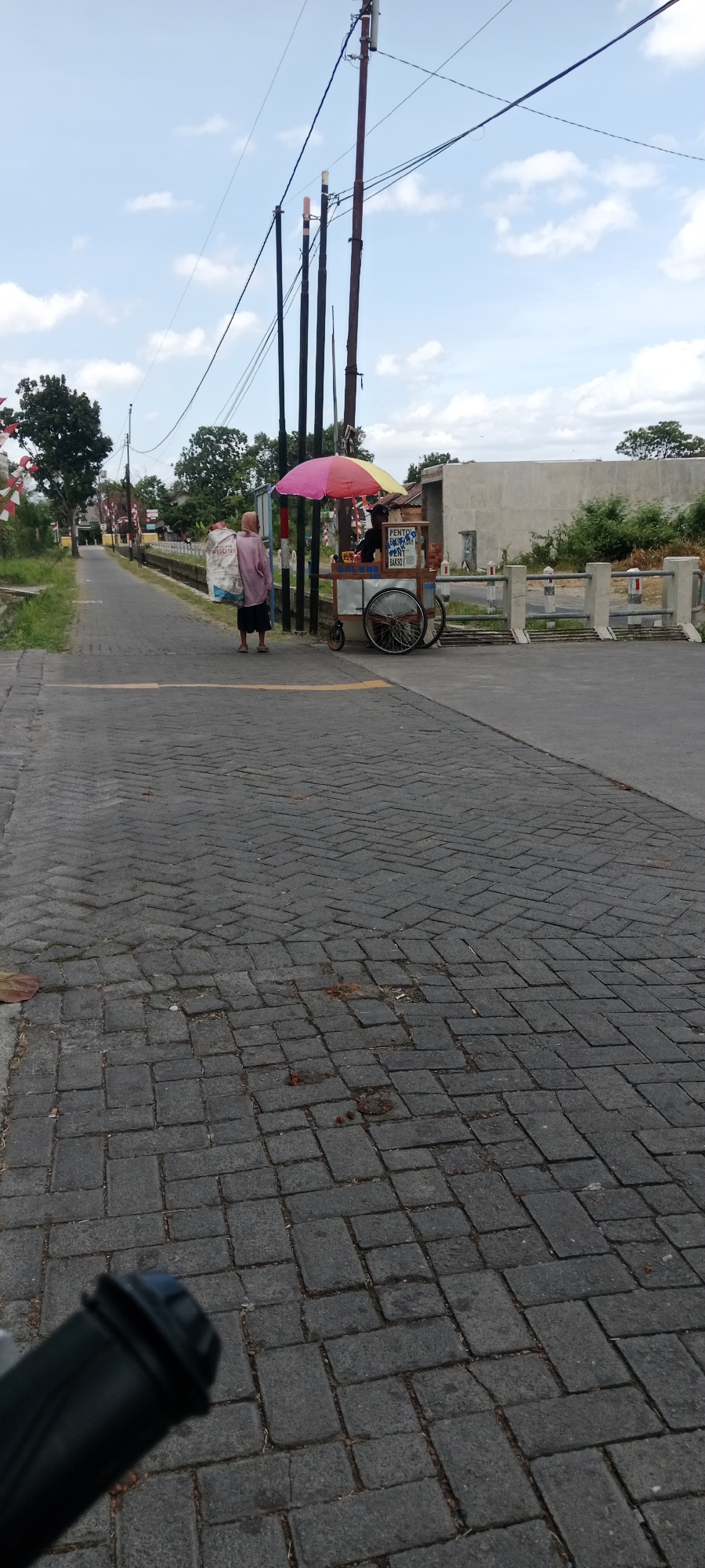 The image depicts a street scene in Maguwoharjo, Indonesia. In the foreground, a dark, textured surface of interlocking pavers suggests a pedestrian walkway or street. The midground features a person in a pinkish-purple garment walking away from the camera, carrying a large, white bag over their shoulder. To the right of the pedestrian is a food cart with a multi-colored umbrella. The cart has wheels and appears to be selling food, with some text visible on its side. A vendor is standing behind the cart. In the background, there are utility poles with wires, trees, a concrete wall, and a barrier with vertical posts, indicating a transition point or an entrance. The sky is partly cloudy. The overall setting is outdoor, likely in a moderately populated area.