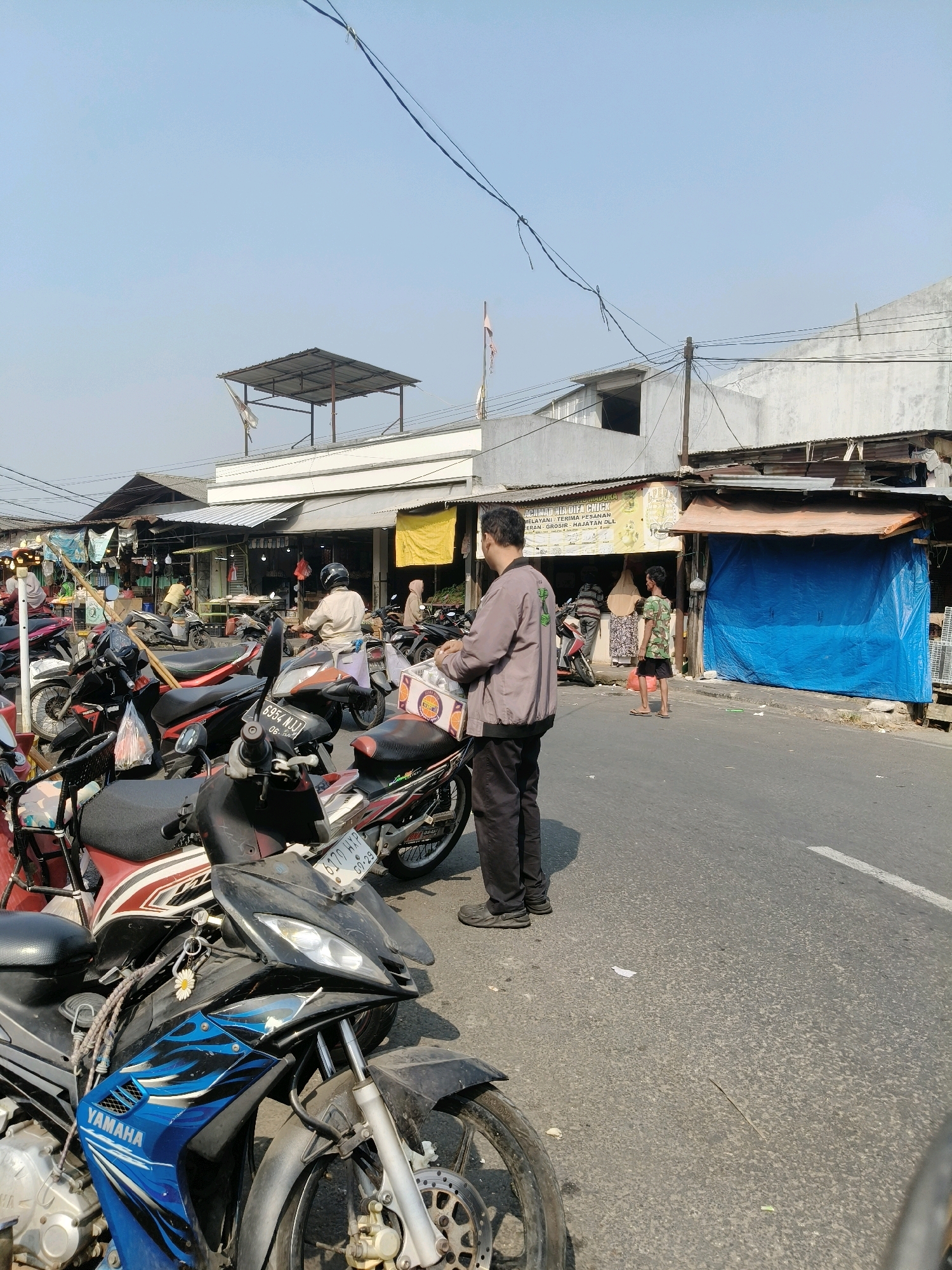 A daytime, outdoor scene captures a street market in South Tangerang, Indonesia. A man in a grey jacket and black pants stands facing away from the camera, holding a white box. He is on a paved road with several motorcycles parked to his left. In the background, a row of market stalls with corrugated metal roofs and awnings line the street. One stall is covered with a blue tarp. Several people are visible in the market, some standing and some on motorcycles. The sky is clear and blue.
