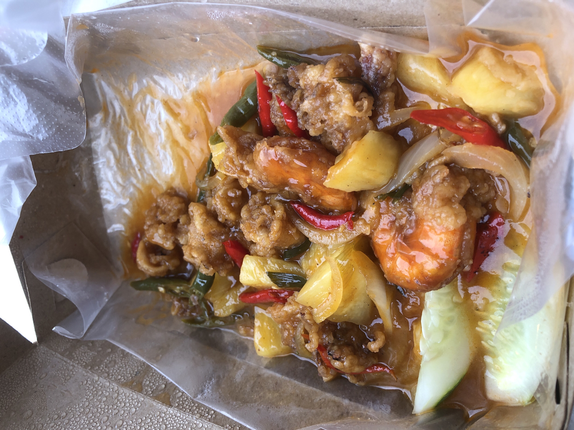 The image shows a takeout container filled with food. The food appears to be a dish with fried shrimp, pineapple chunks, onion slices, red and green peppers, and slices of cucumber, all coated in a sauce. The food is contained within a clear plastic bag or liner placed inside the takeout container. The takeout container is sitting on a surface that appears to be textured. The location is Unknown, Unknown.