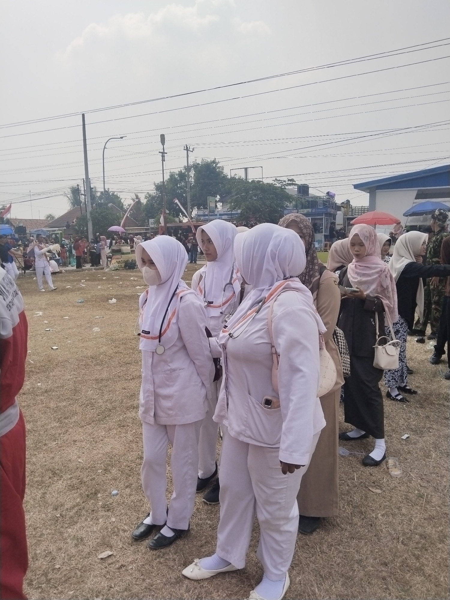 In Margasari, Indonesia, a group of individuals, primarily women wearing hijabs and light-colored uniforms, stand in an outdoor area with dry, grassy ground. Two women in the foreground are wearing white scrub suits, white hijabs, and have stethoscopes around their necks. One of them is wearing a face mask. The woman to the left has a phone clipped to her scrub top pocket. Other women in the background are also wearing hijabs of various colors and patterns, some in casual attire and others in darker uniforms. There are tents and structures in the background, suggesting an event or gathering. Power lines and utility poles are visible overhead against a cloudy sky. The ground is littered with some small pieces of debris.