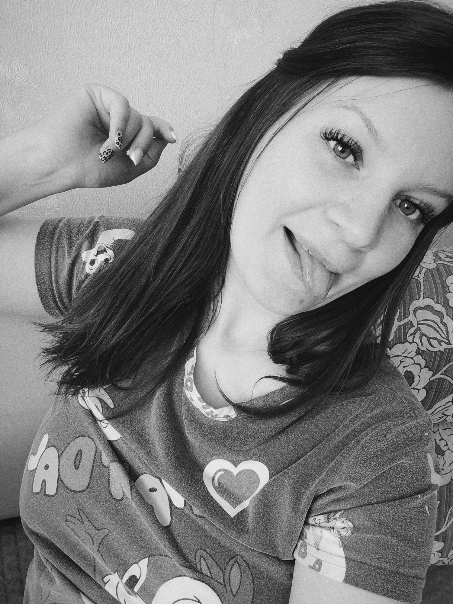 A woman takes a selfie indoors. She has long dark hair and is wearing a t-shirt with a cartoon character on it. Her nails are decorated with a black and white animal print. She is sticking out her tongue. The environment includes a patterned cushion and a textured wall. It's impossible to discern if the location is Tobolsk, Russia from the image provided.