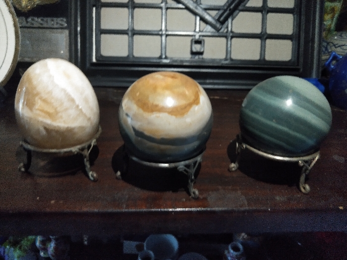 The scene, located indoors in Steenbergen, Netherlands, displays three polished mineral spheres arranged horizontally on a dark, polished wooden surface. From left to right: The first sphere, appearing ovoid, is light brown and white with swirling patterns, supported by an ornate, tarnished silver-colored metal tripod stand. The central sphere is spherical, displaying concentric bands of tan, white, and dark grey-blue, resting on a black circular base which is itself supported by a simpler dark metal tripod stand. The third sphere is spherical, featuring horizontal bands of dark teal and lighter green, and sits on an ornate, tarnished silver-colored metal tripod stand matching the first. The background is out of focus, revealing a dark rectangular grid structure behind the spheres. To the far left, a partial round white object with a gold rim displays the partial text 'ASSES'. To the far right, a partial blue ceramic object is visible. No individuals are present, and the scene is static.